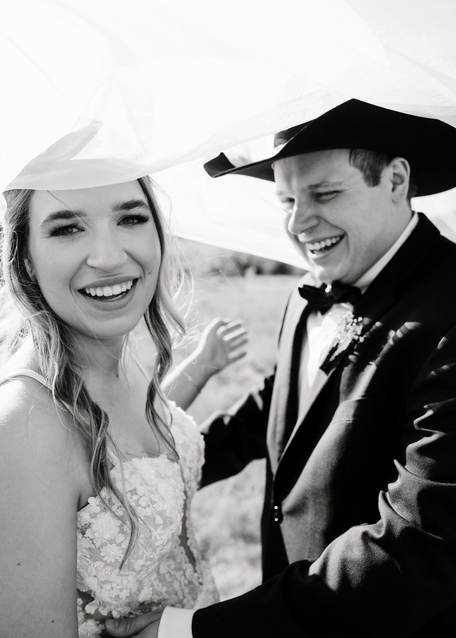 Black and white photo of a smiling bride in lace dress and groom in cowboy hat laughing together on their wedding day.