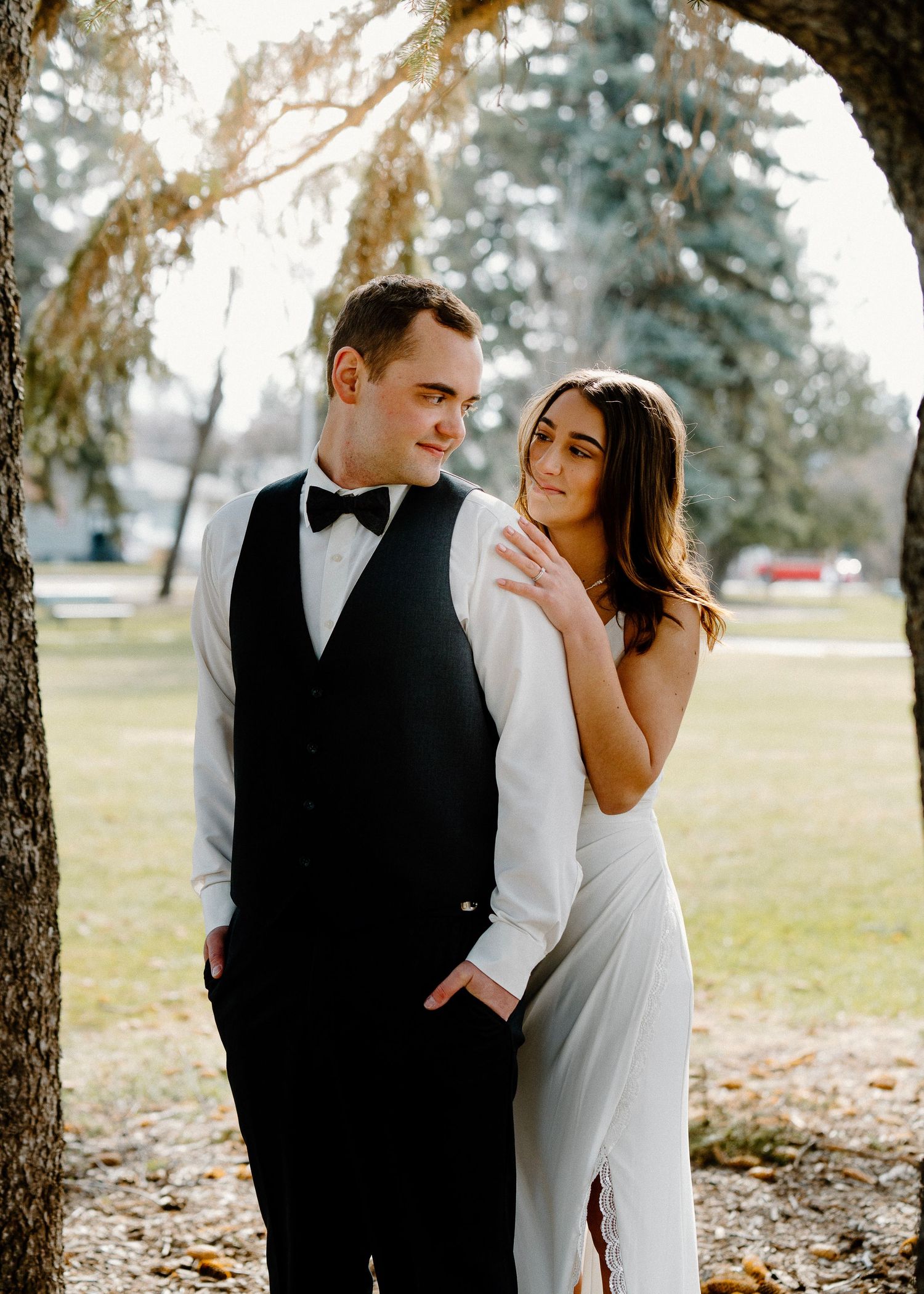 Elegant couple in wedding attire posing under a tree outdoors, groom in black tuxedo vest and bride in white gown.