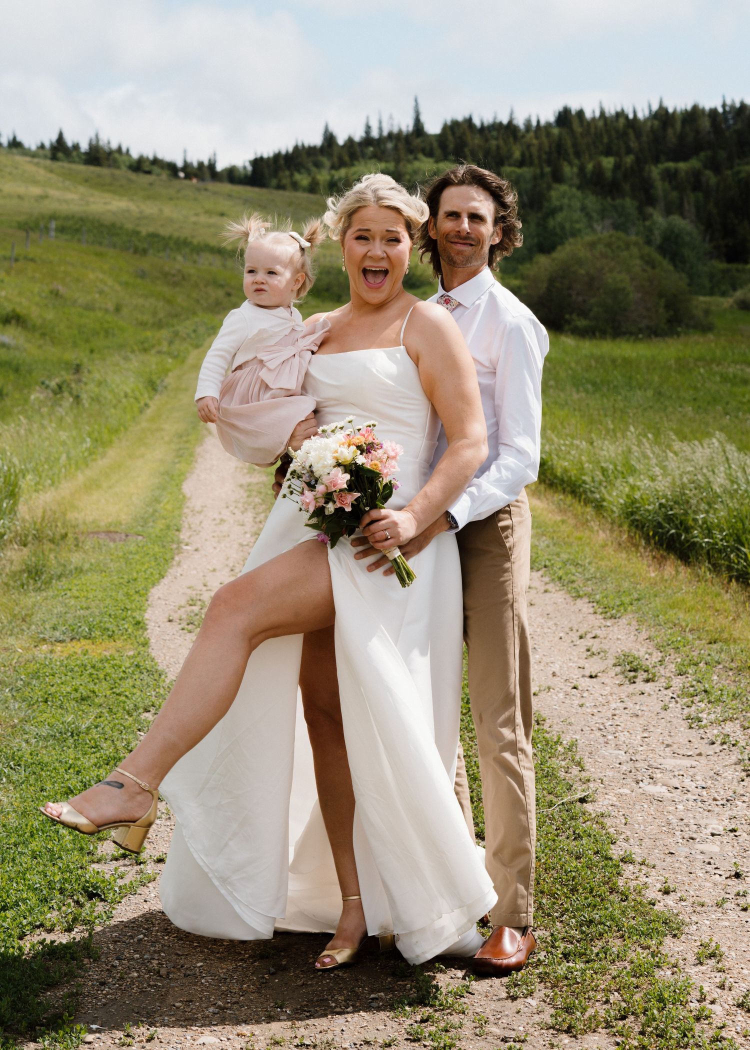 Happy wedding couple with baby posing on a scenic dirt path surrounded by green hills.