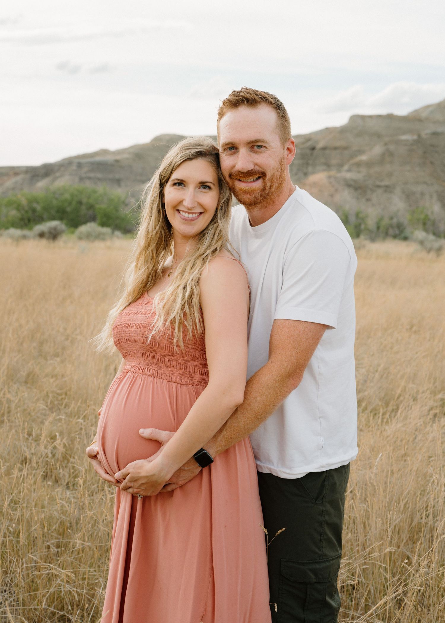 Smiling couple in a golden field, pregnant woman in coral dress held by partner.