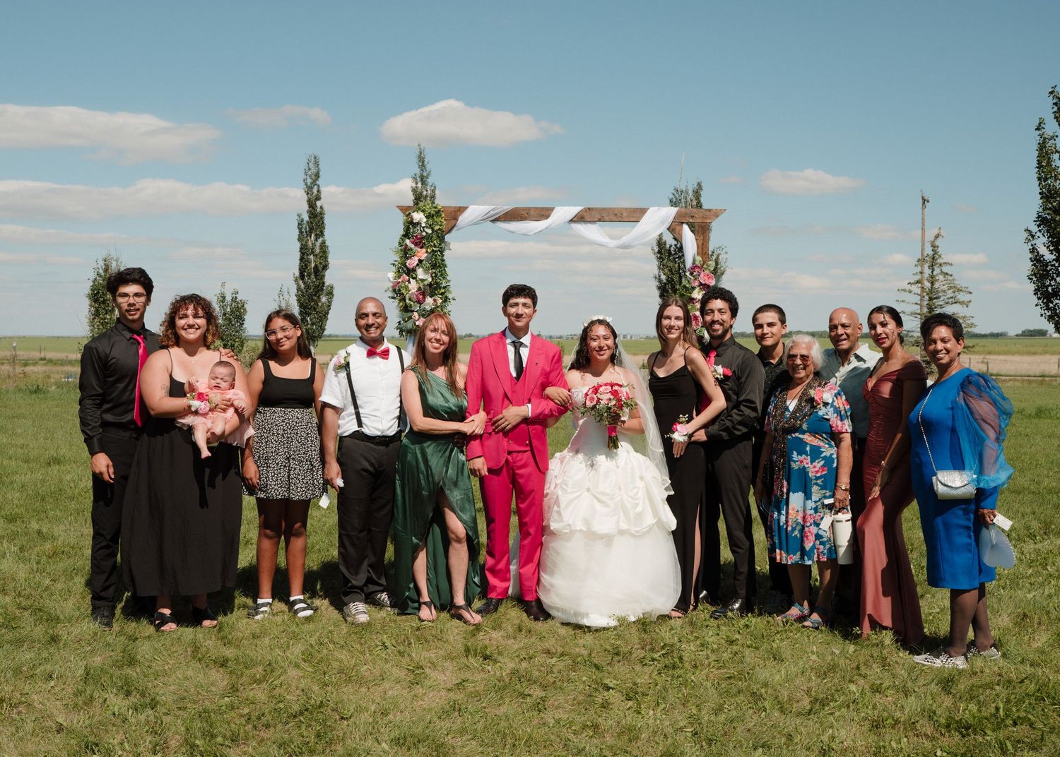 Wedding party group photo outdoors under floral arch on sunny day in open field.