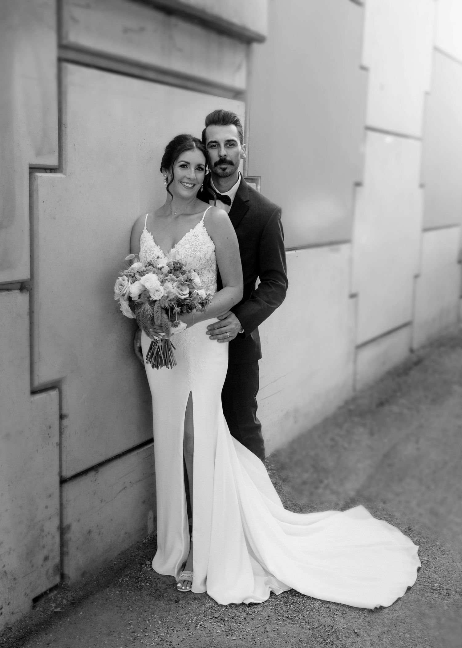 Elegant bride and groom pose in black and white portrait against a stone wall with bouquet.