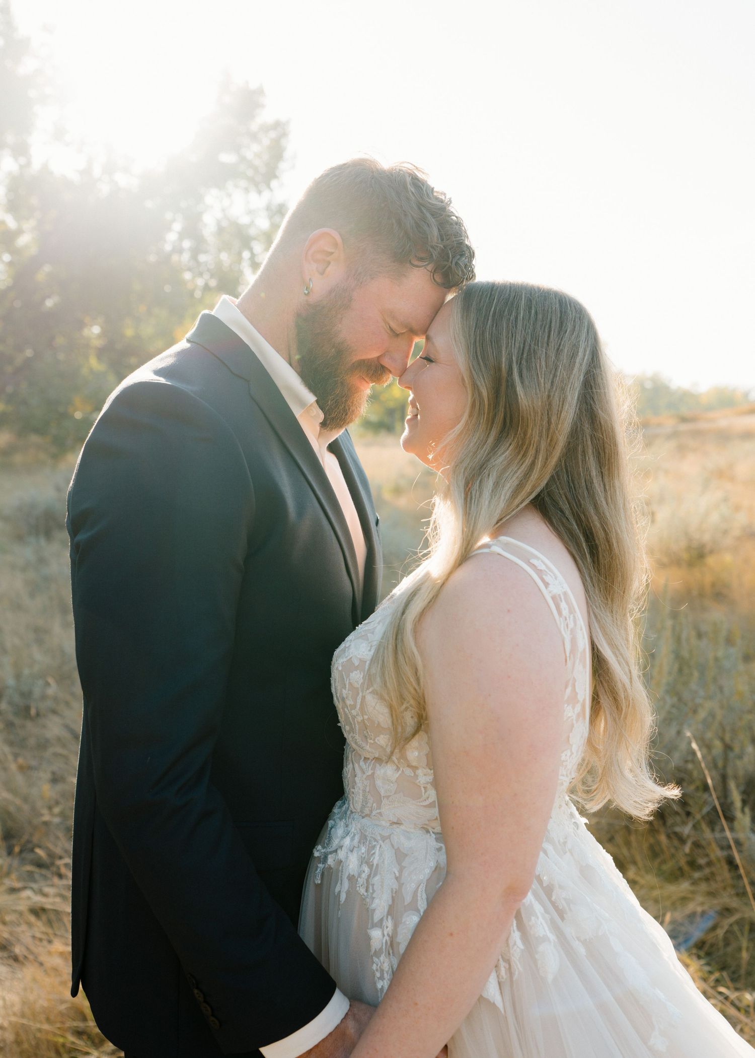 Bride and bearded groom touching foreheads in a tender moment with golden sunlight glowing behind them.