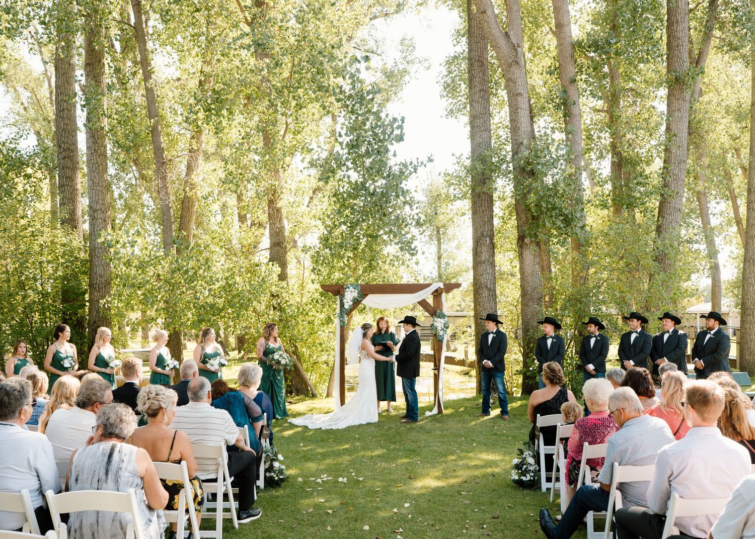 Outdoor wedding ceremony in wooded setting with guests seated in white chairs and wedding party at altar.