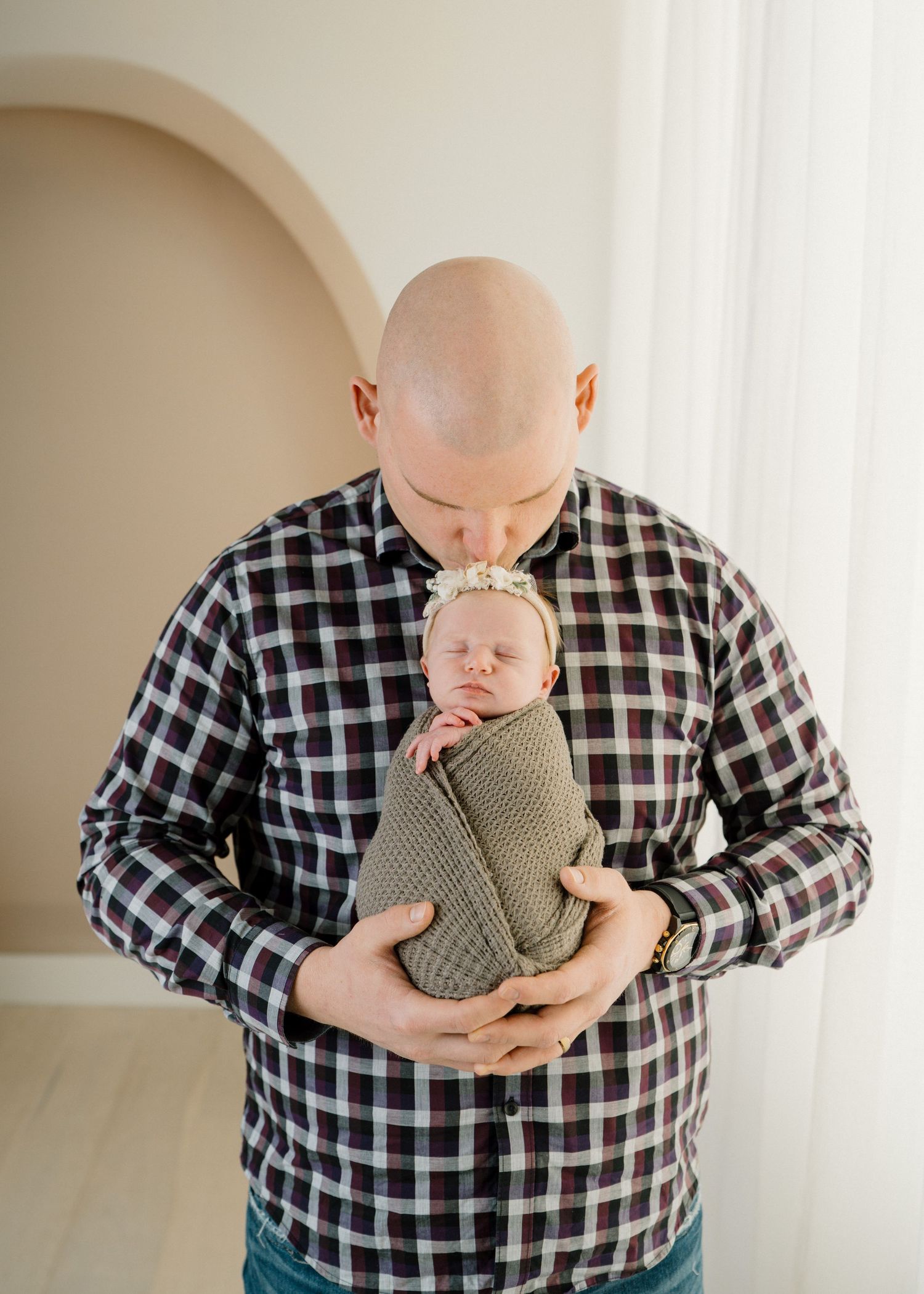 Bald father in plaid shirt tenderly kisses swaddled newborn baby with floral headband.