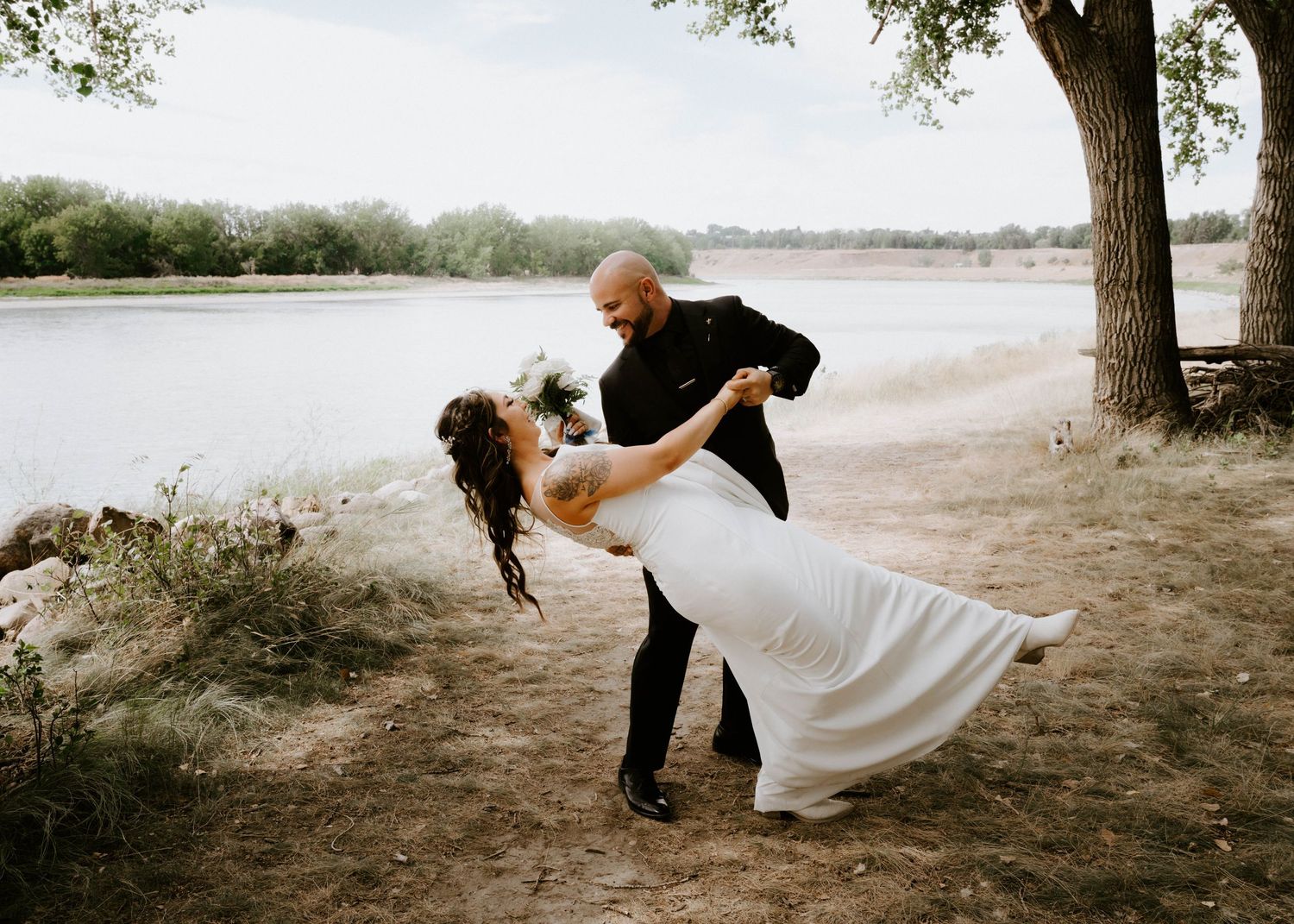 Groom dips bride in white wedding dress near a lake under a large tree.