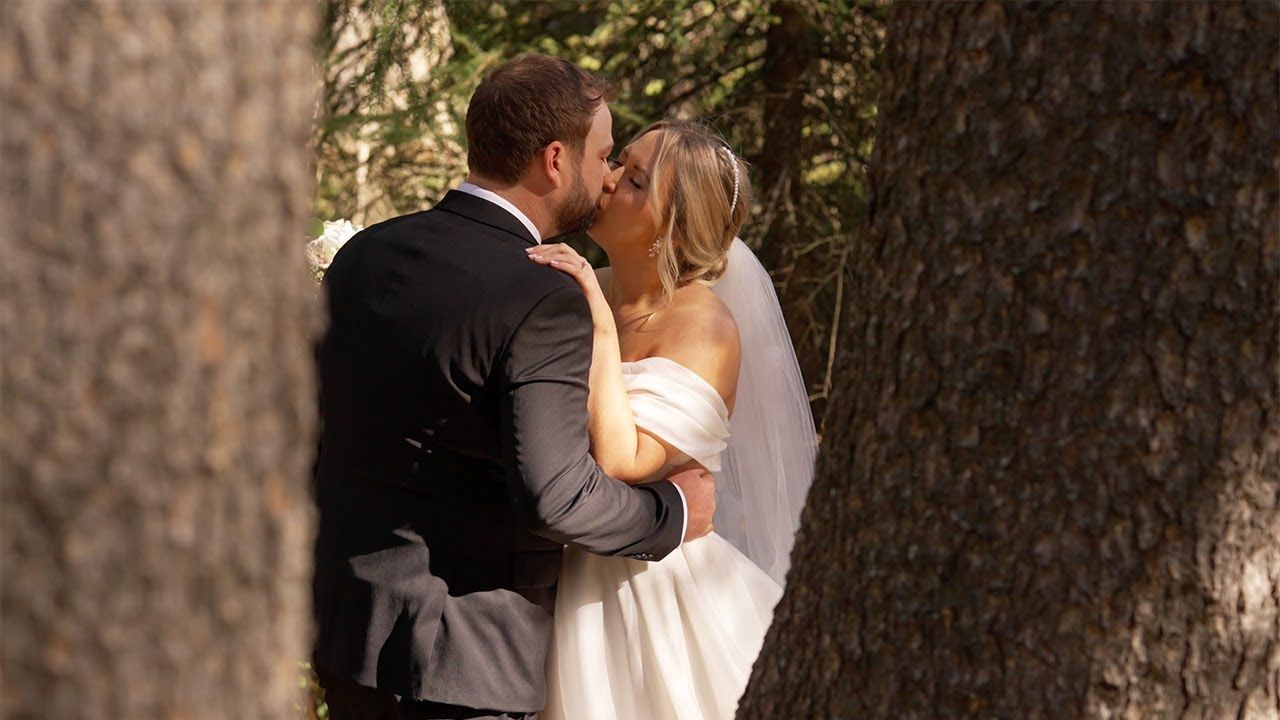 A bride and groom share a romantic kiss outdoors near trees, with the bride wearing a white gown and veil.