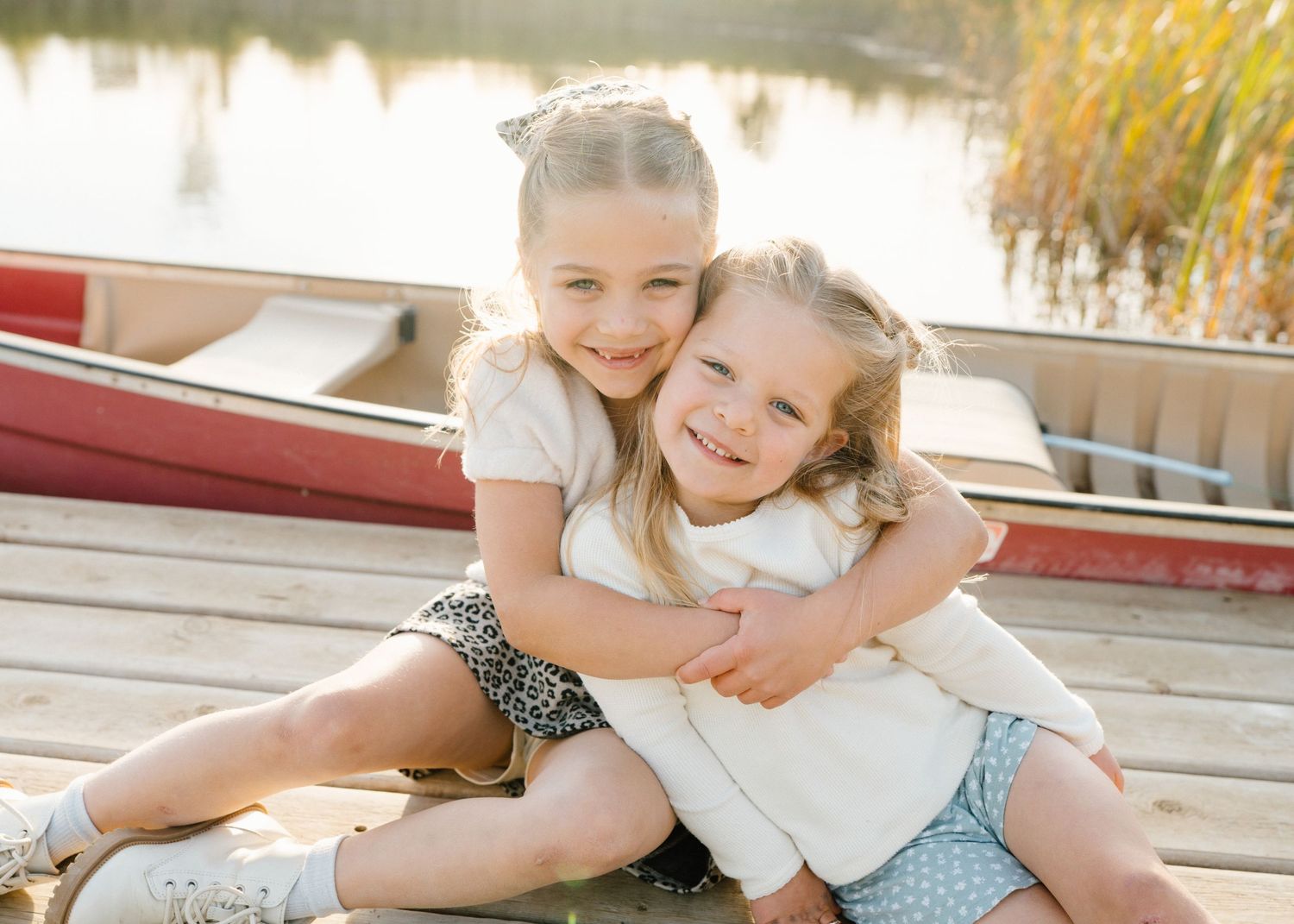 Two young sisters hugging and smiling on a wooden dock near a red canoe on a sunny day.