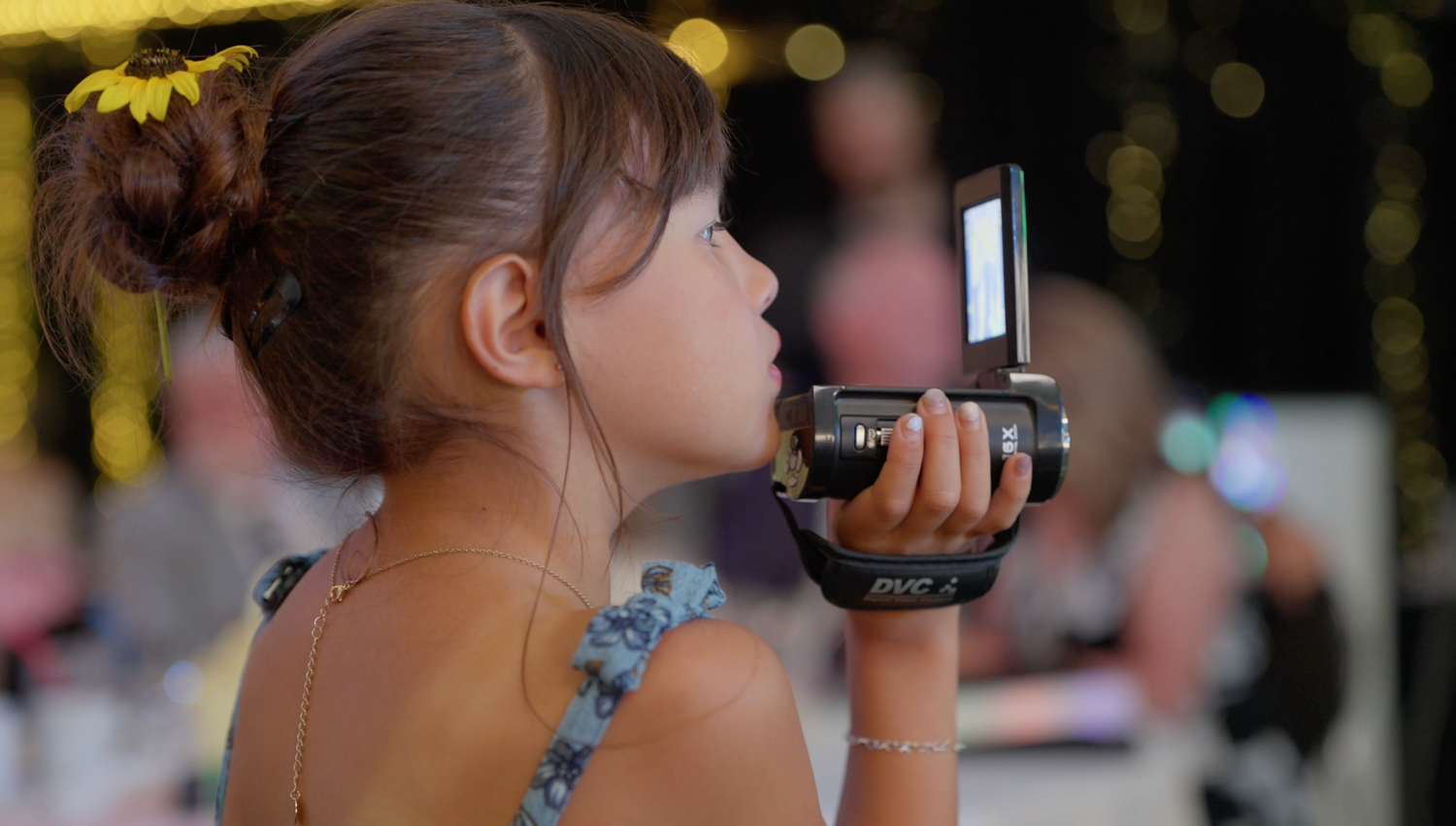 A young girl with a flower in her hair holds a vintage camera up to photograph at an outdoor event.