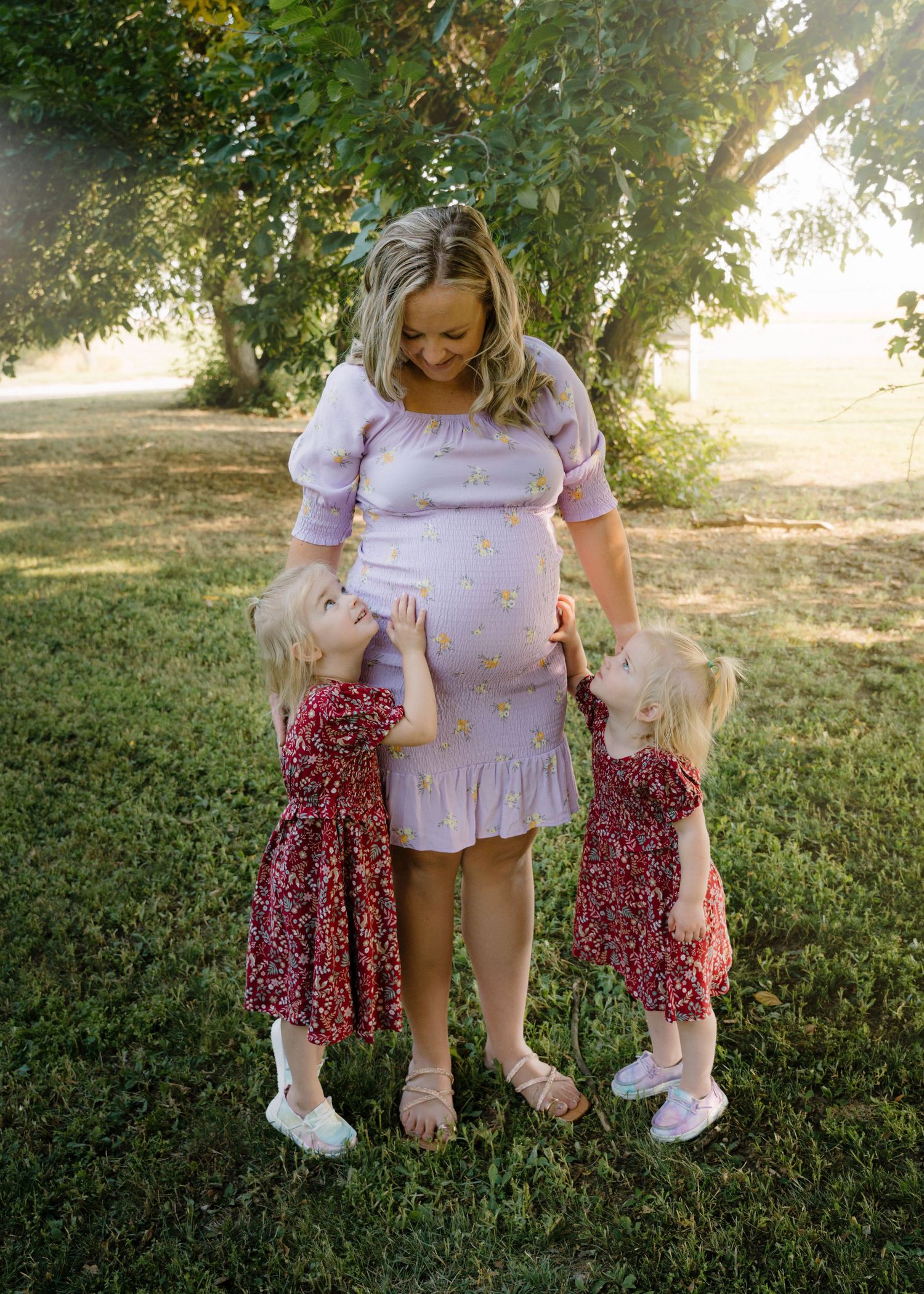 Pregnant woman in lavender dress smiling at two young daughters in red floral dresses outdoors.