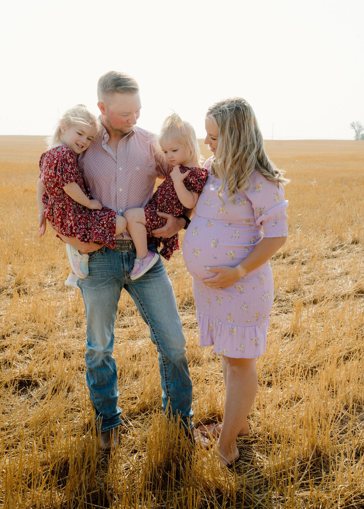 Father holding two daughters in red dresses while pregnant wife in lavender dress looks on in field.