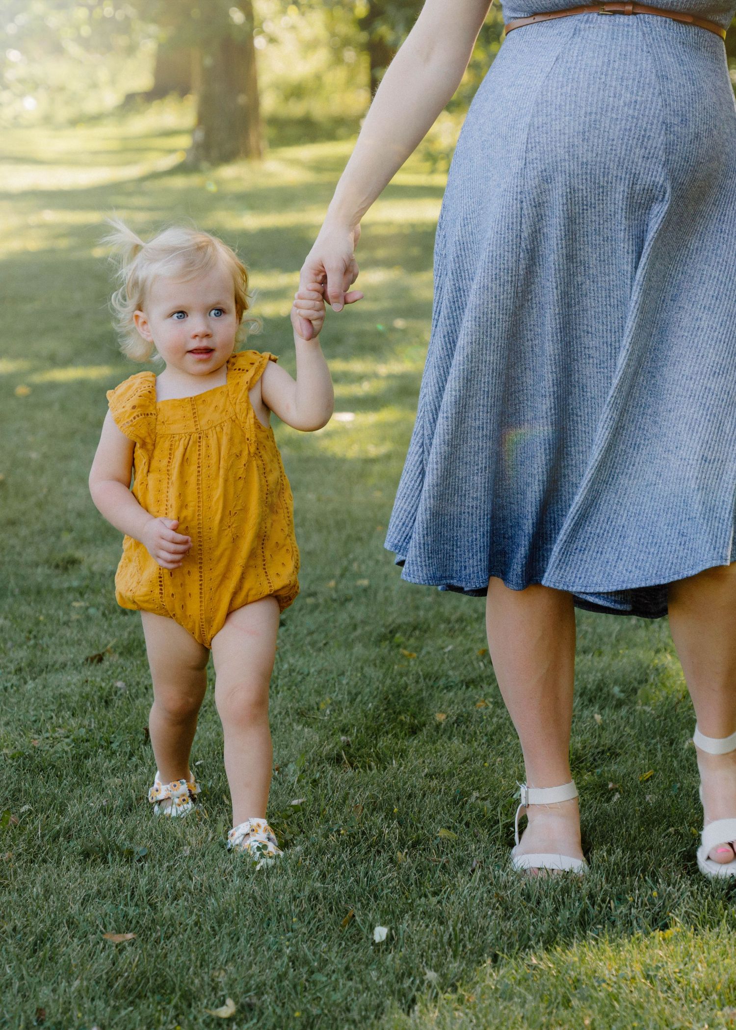 Adorable blonde toddler in mustard yellow romper holding mothers hand while walking on lush green grass outdoors.