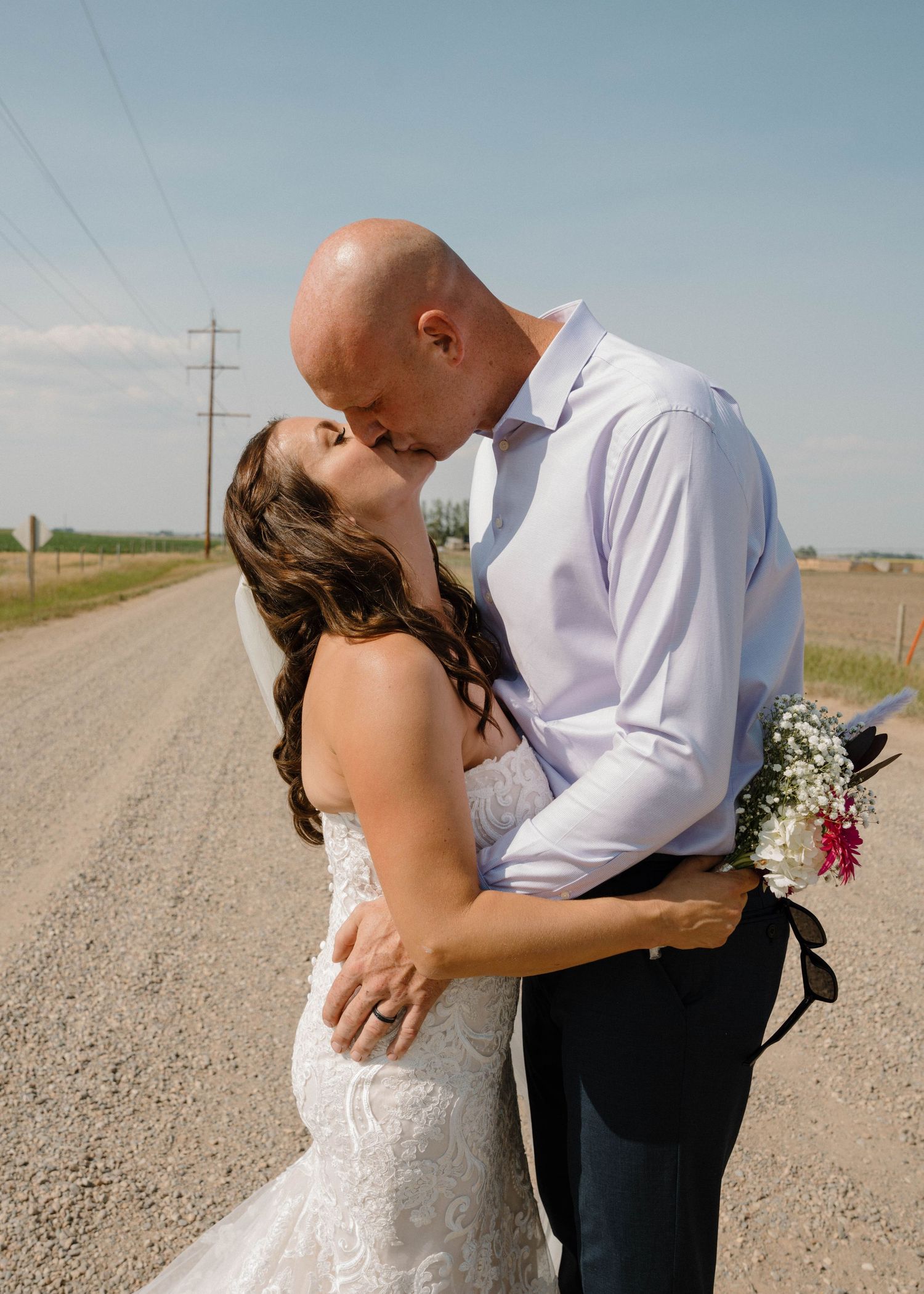 Bride and groom share a romantic kiss on a sunny rural dirt road during wedding photos.