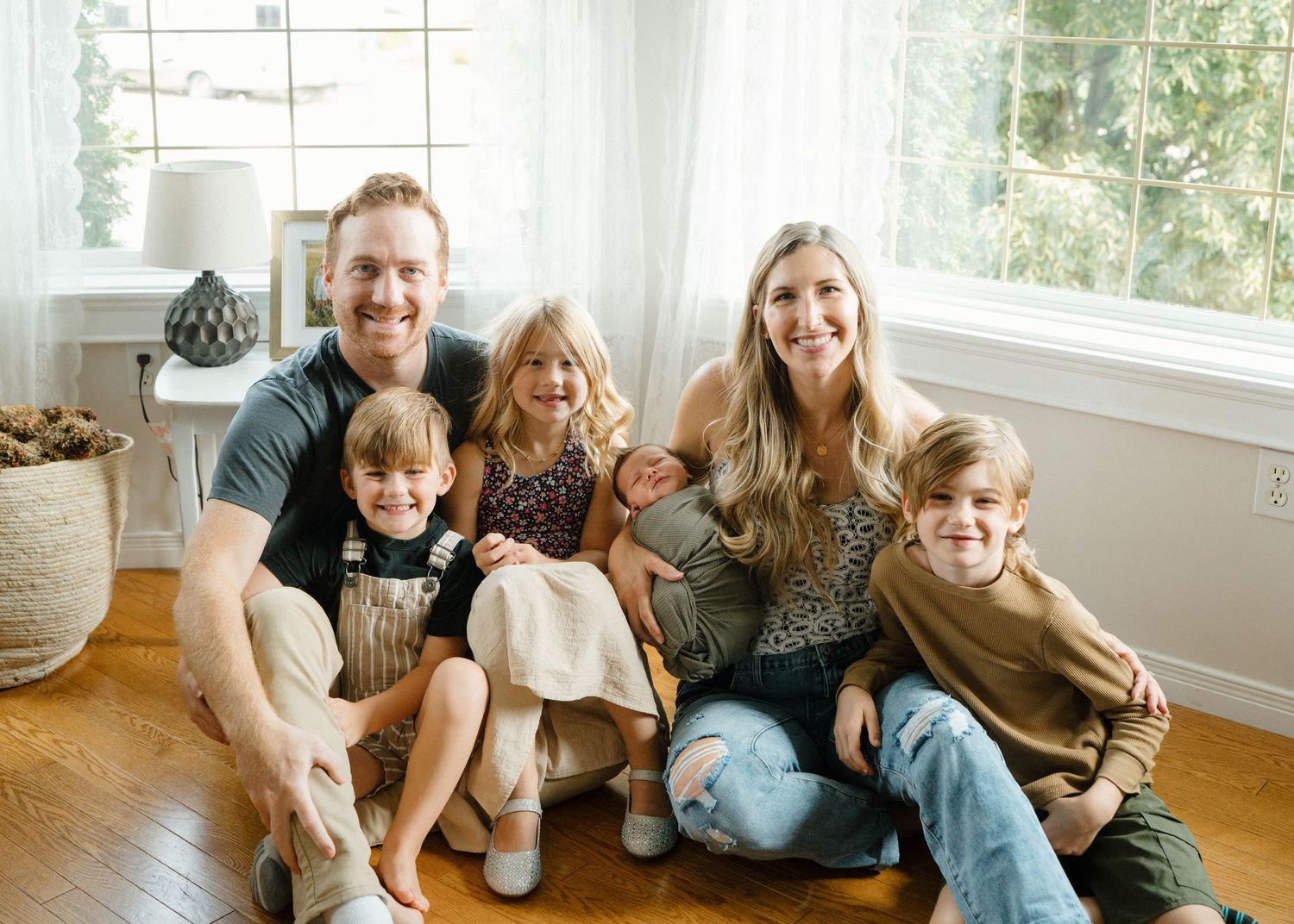 Smiling family of six with newborn baby sits together on hardwood floor in bright indoor setting.