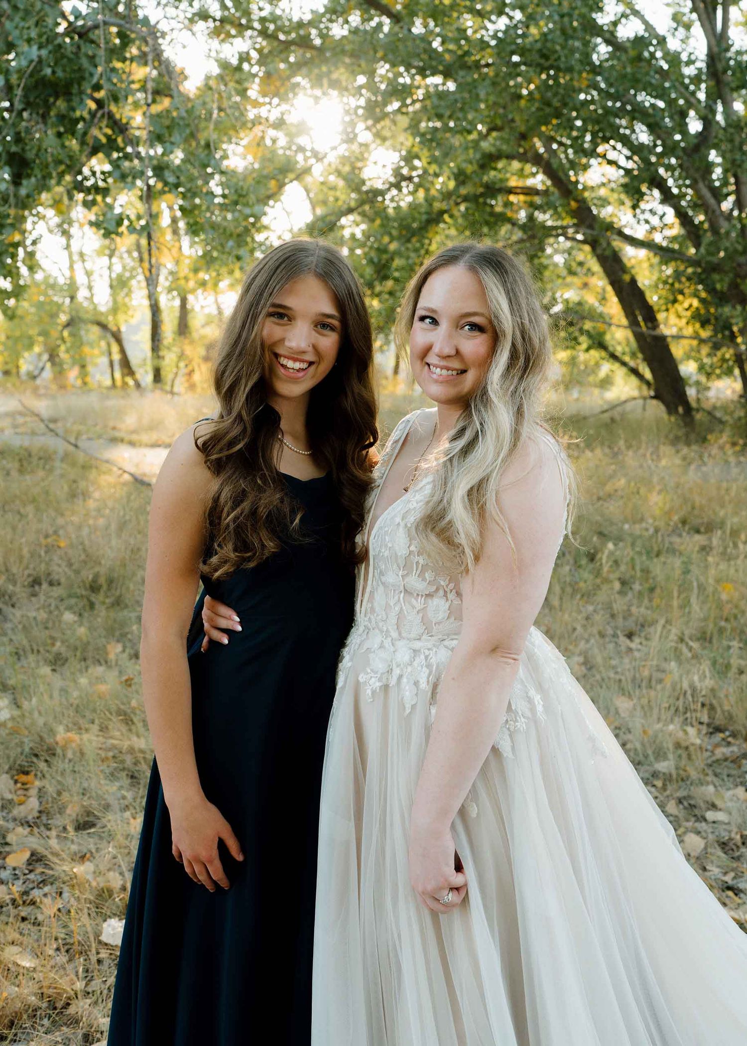Two young women in formal gowns, one black and one white lace, posing outdoors among golden trees.