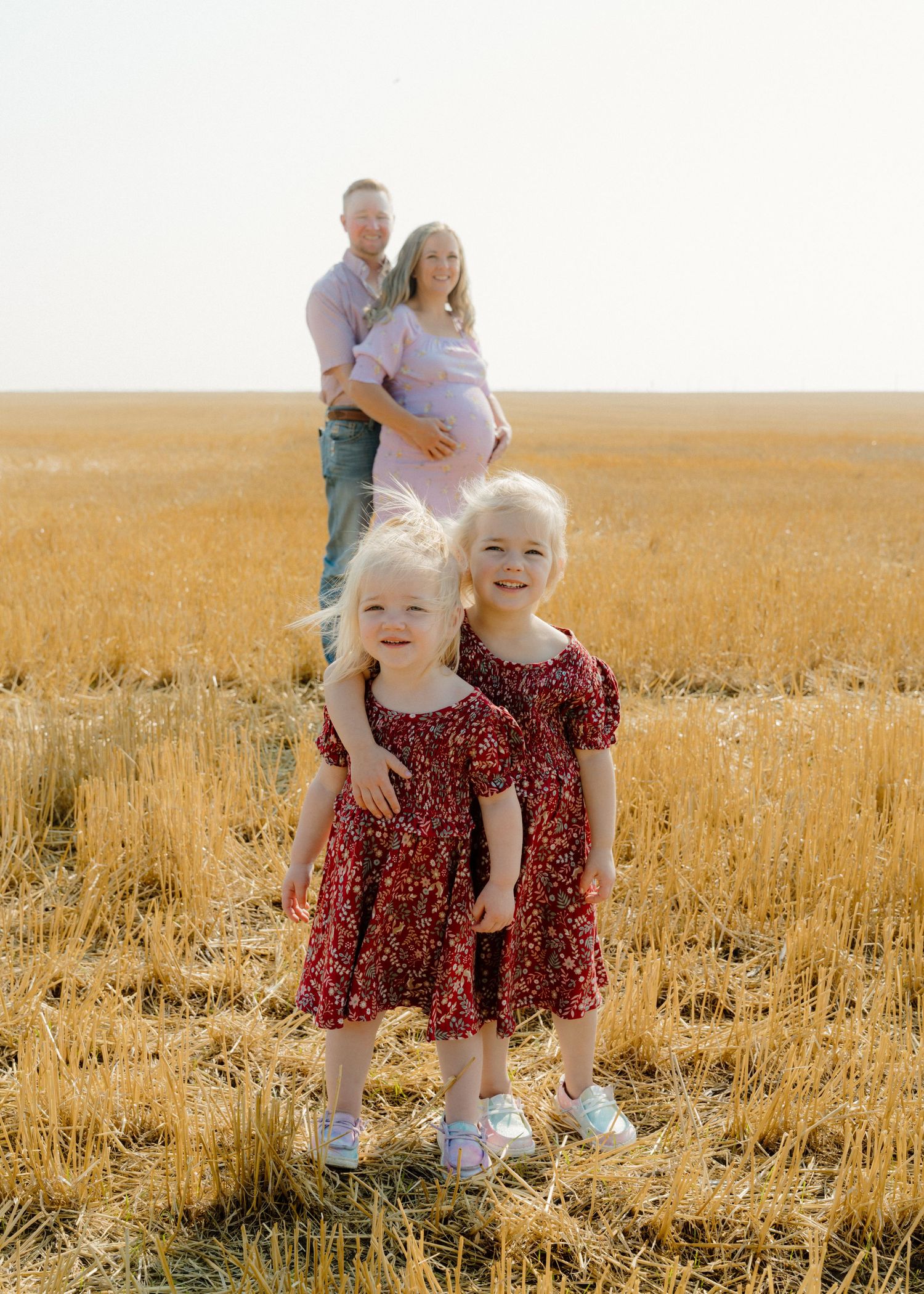 Family maternity photo with two young girls in red dresses posing in golden wheat field.