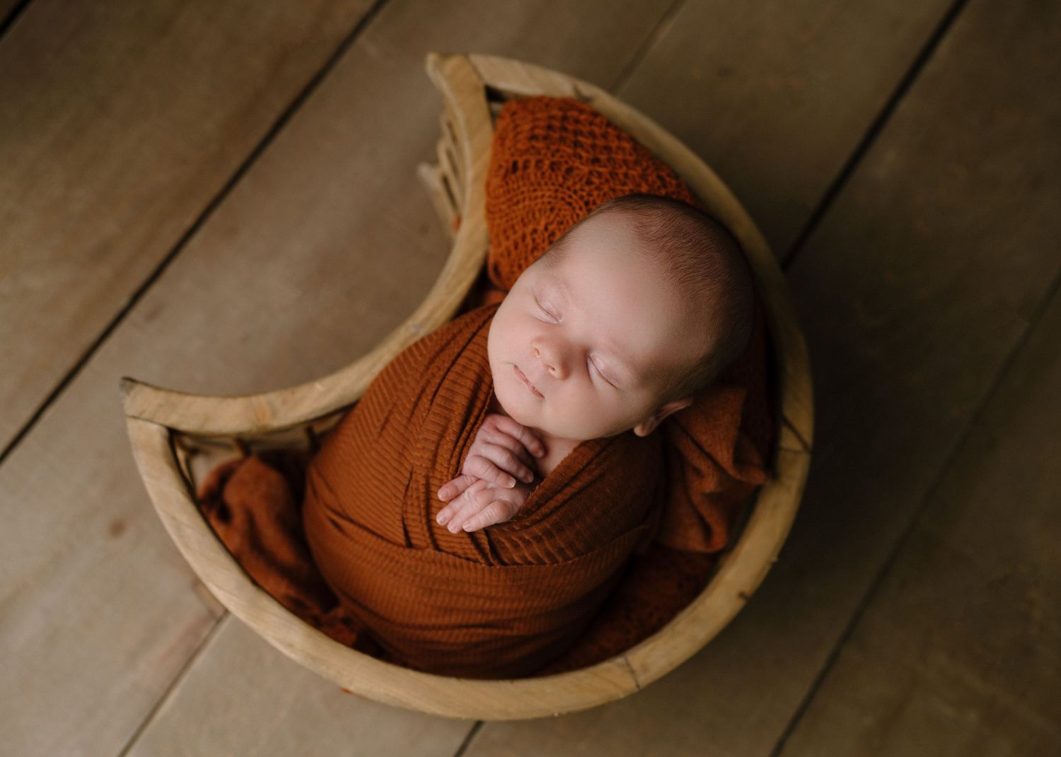 Sleeping newborn baby wrapped in burnt orange swaddle and knit bonnet posed in a crescent moon wooden bowl.
