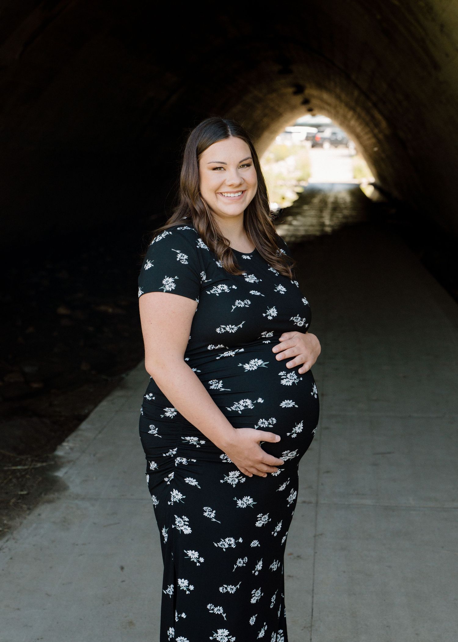 Pregnant woman in black floral dress poses confidently inside a rustic stone tunnel walkway.
