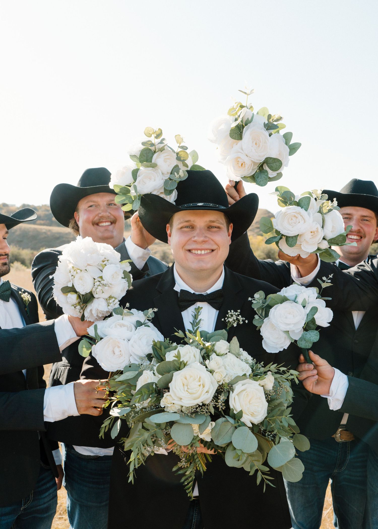 Smiling groom surrounded by groomsmen in cowboy hats holding white rose and eucalyptus bouquets.