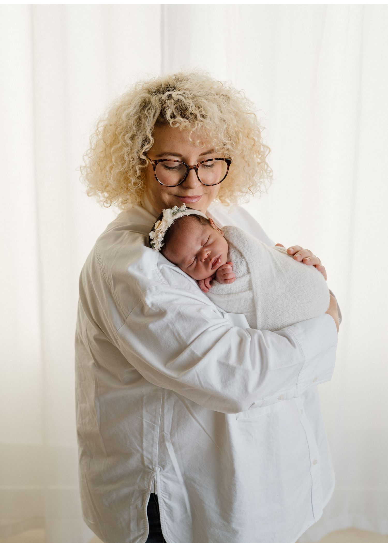 Curly blonde mother with glasses tenderly holding sleeping swaddled newborn against white curtains.