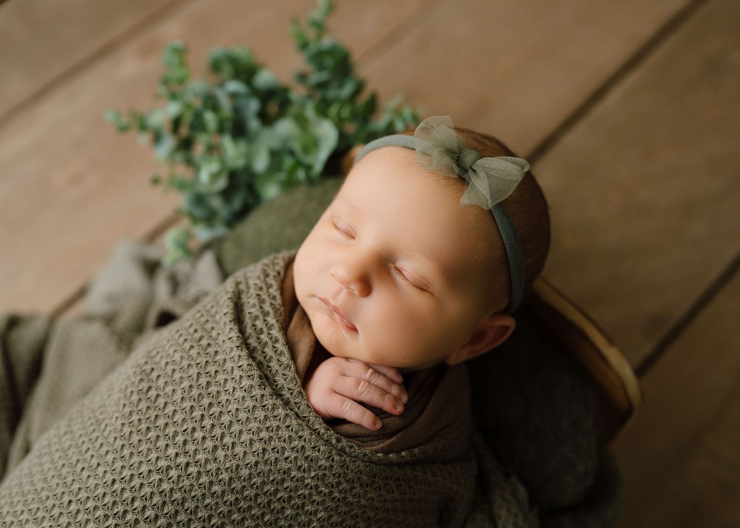 Sleeping newborn baby wrapped in olive green swaddle with matching bow headband in a wooden bowl.