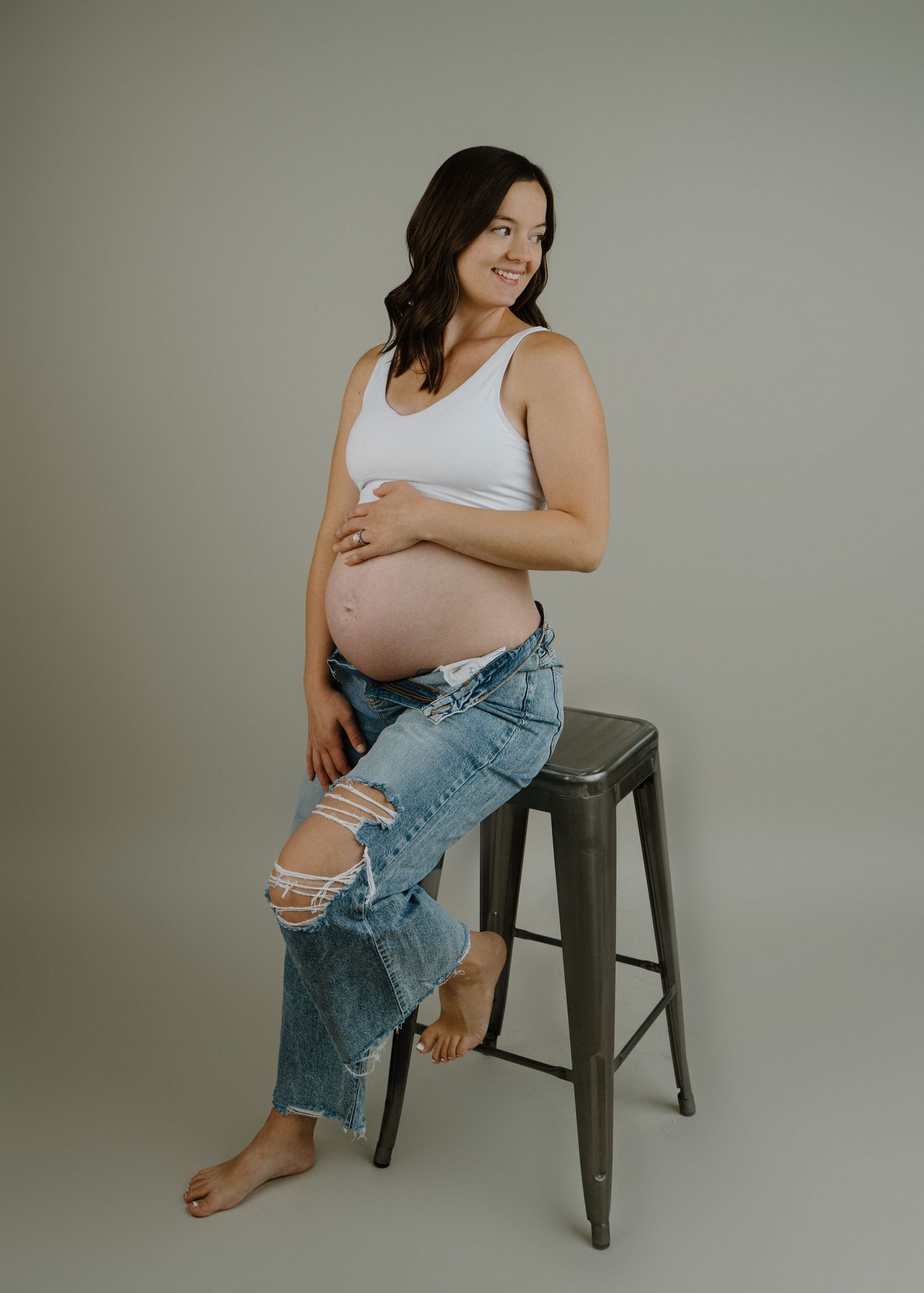Smiling pregnant woman in white tank and ripped jeans poses beside a metal stool in studio.