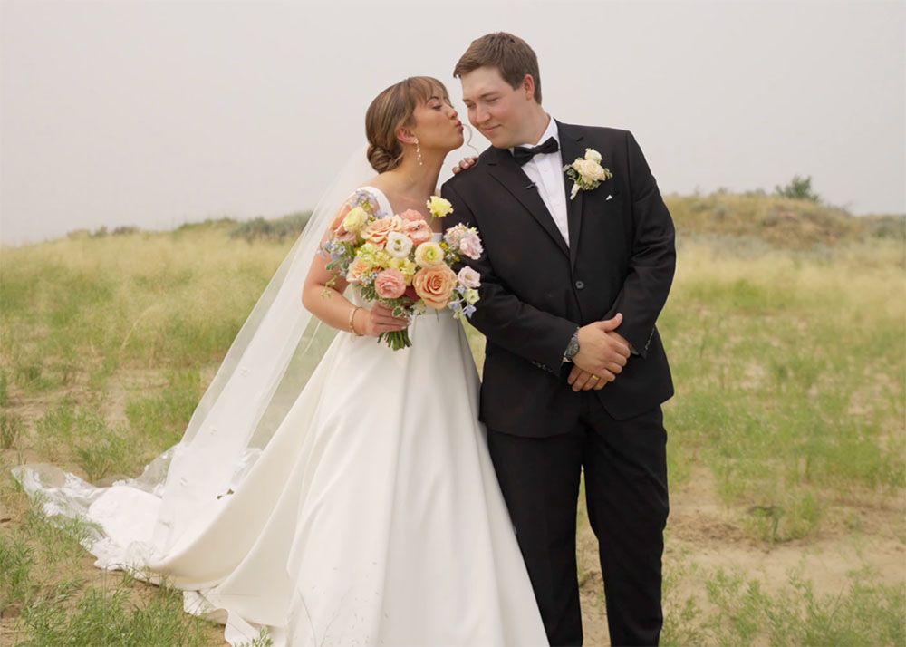 Bride in white gown kisses groom in black tuxedo holding pink bouquet outdoors in grassy field.