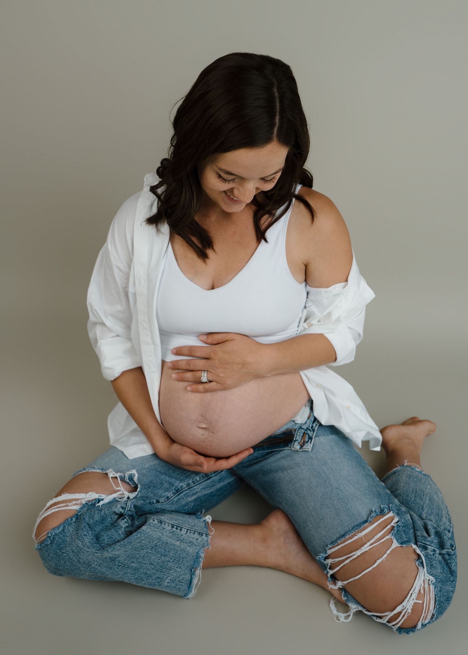 Pregnant woman in white top and ripped jeans sitting cross-legged cradling her bare baby bump in a studio.