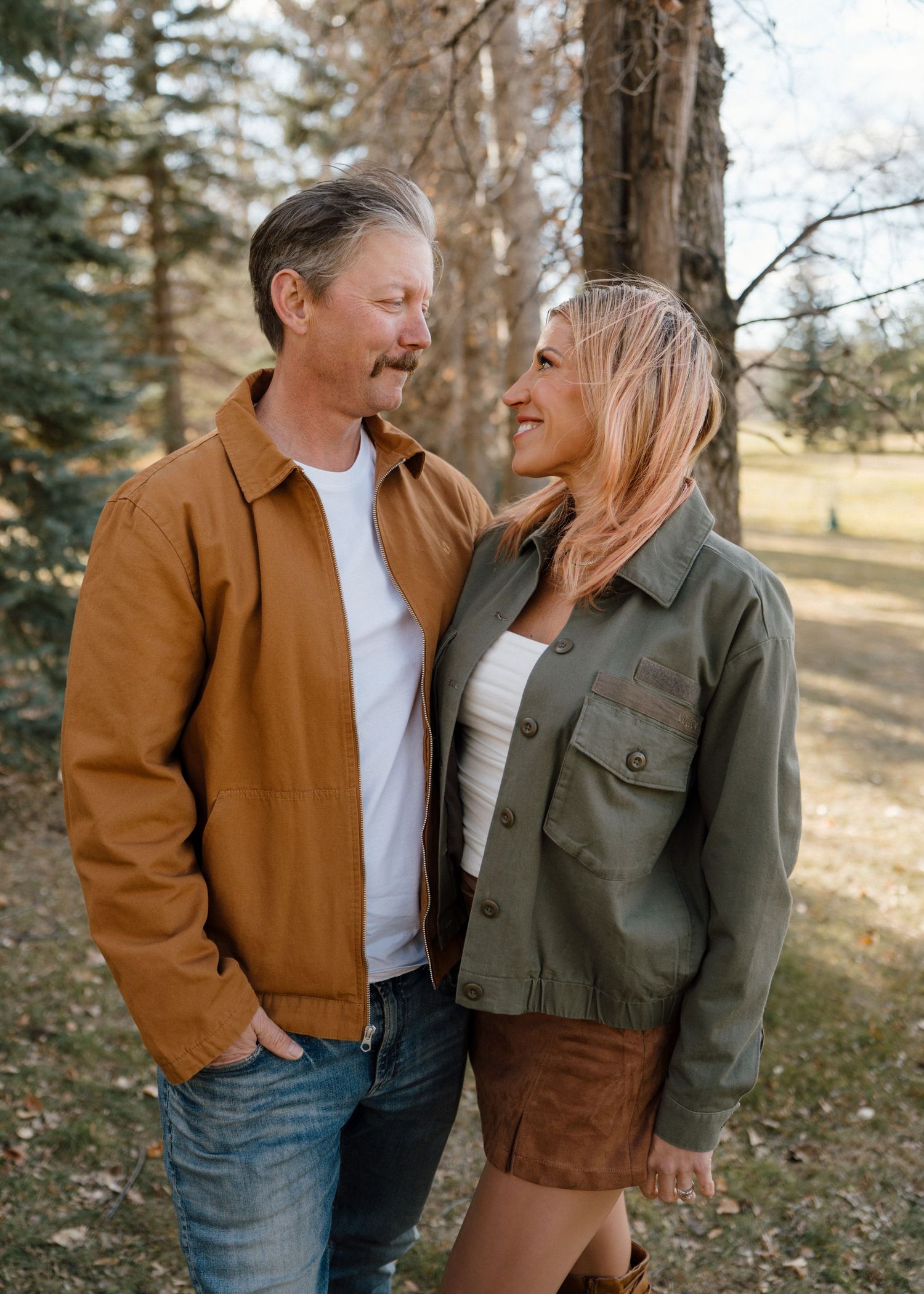 Couple sharing a romantic moment outdoors, man in tan jacket and woman in olive jacket smiling at each other among trees.