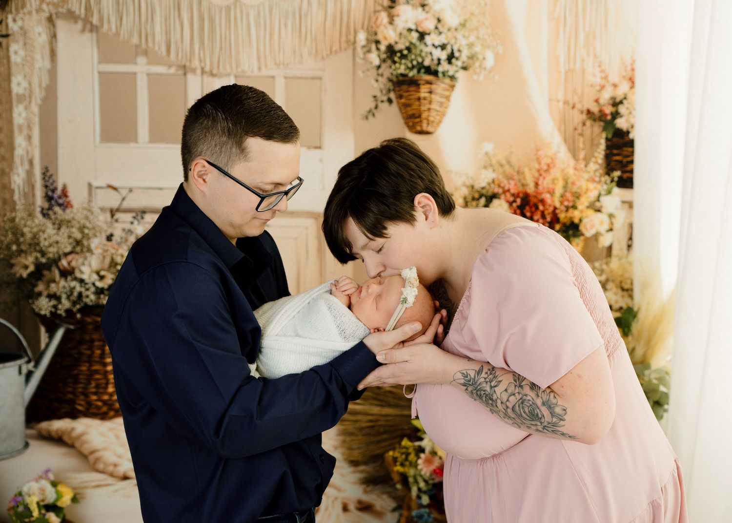 New parents lovingly kiss their newborn baby in a floral decorated photography studio.