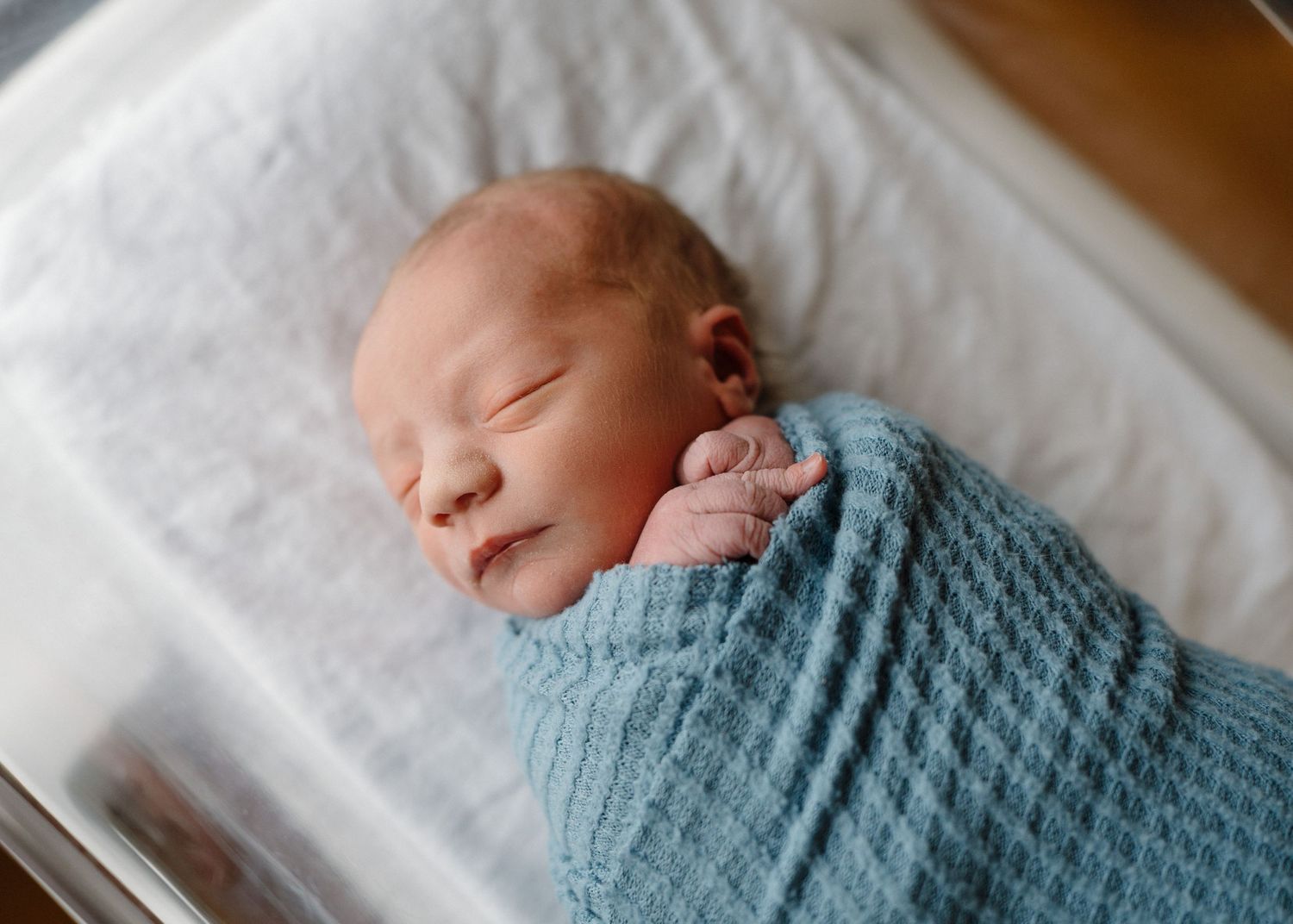 Newborn baby boy swaddled in blue knit wrap sleeping peacefully in white hospital bassinet.