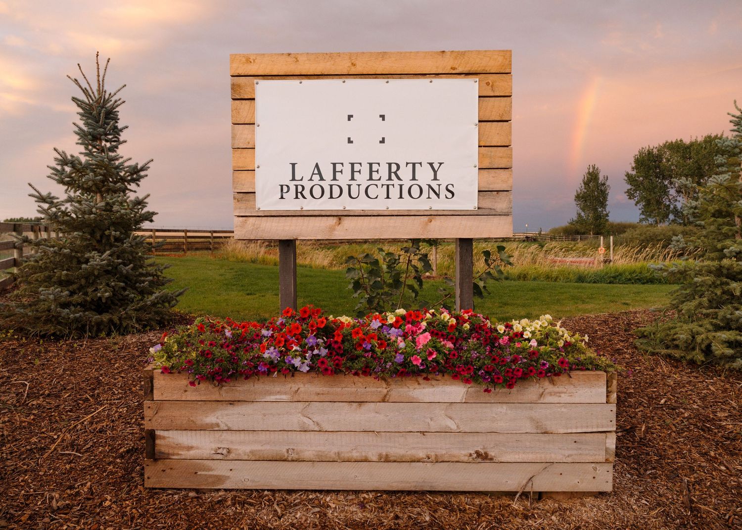 Lafferty Productions wooden sign surrounded by colorful flowers with a rainbow in the background.