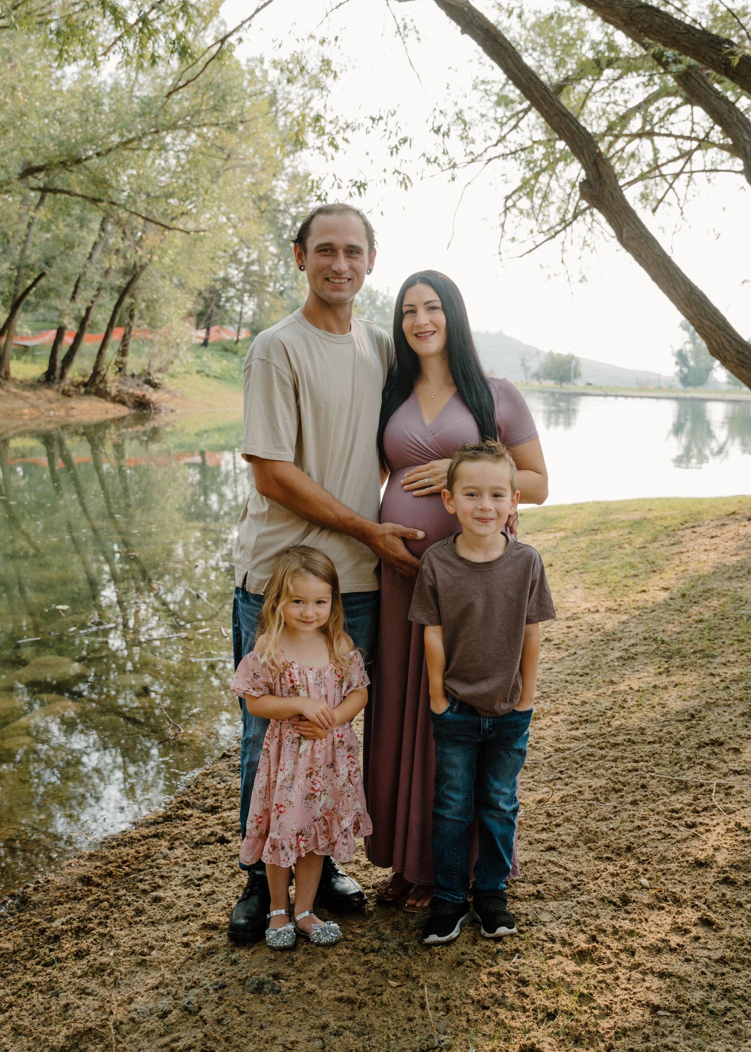 Expecting family of four poses near reflective lake, pregnant mother in purple maxi dress with husband and two children.