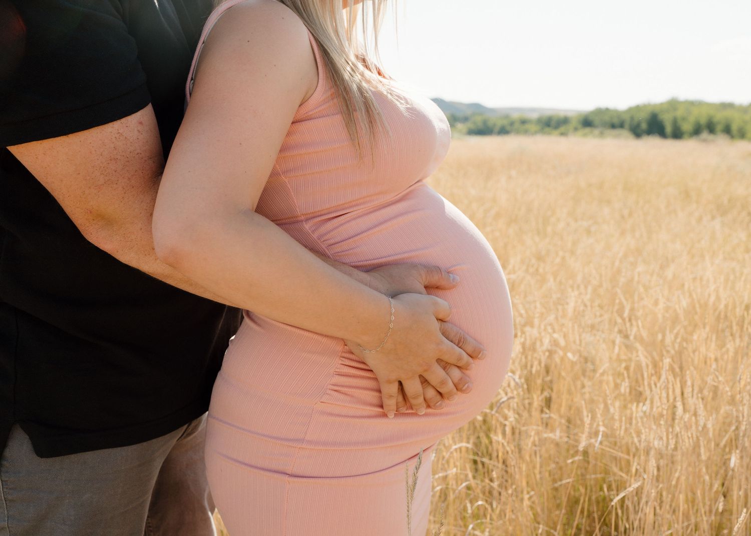 Couple embracing in a sunlit golden field, pregnant woman in a pink dress.