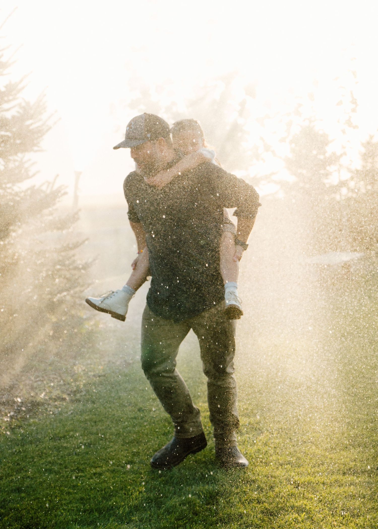 Couple laughing in sprinkler spray on green lawn, man giving piggyback ride in golden sunlight.