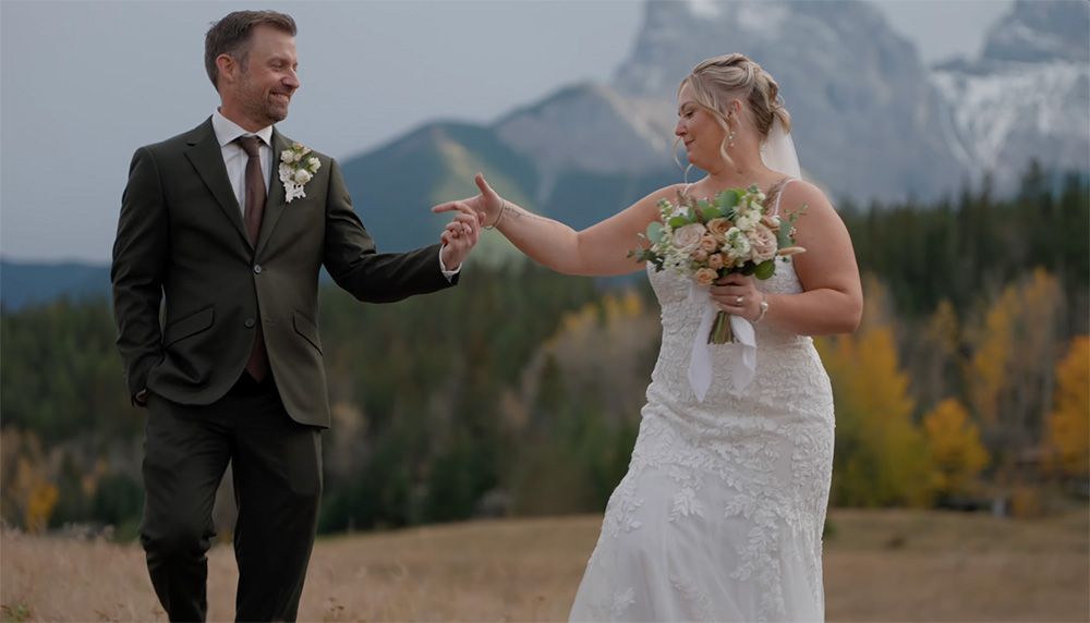 Bride and groom holding hands in a mountain meadow with autumn trees and rocky peaks in the background.