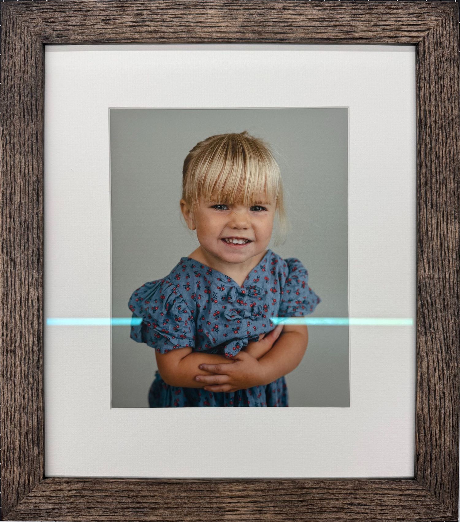 Framed portrait photo of a smiling toddler girl with blonde hair wearing a blue floral dress, arms crossed.