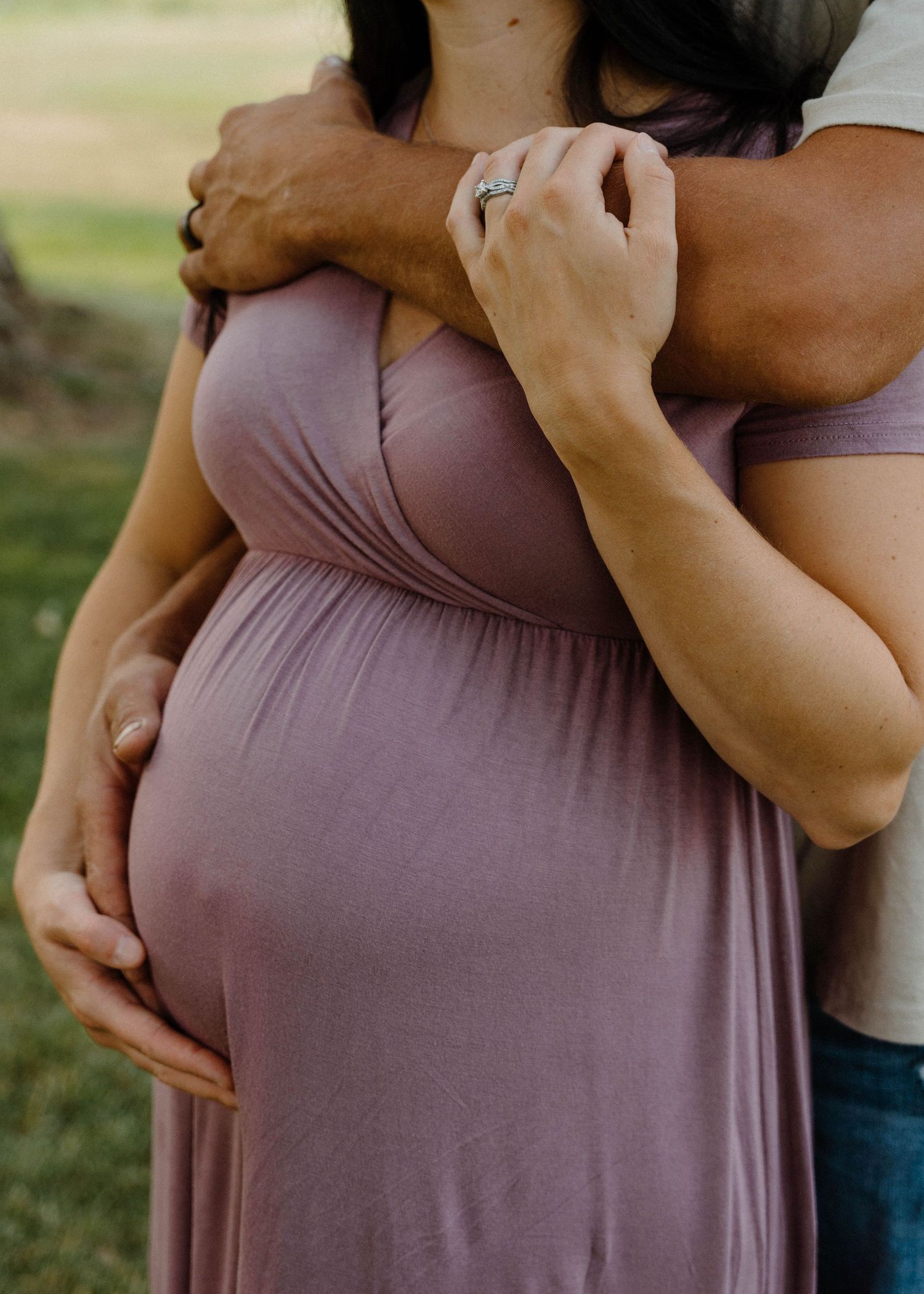 Close-up of pregnant woman in mauve dress with partner embracing her from behind, hands cradling baby bump.