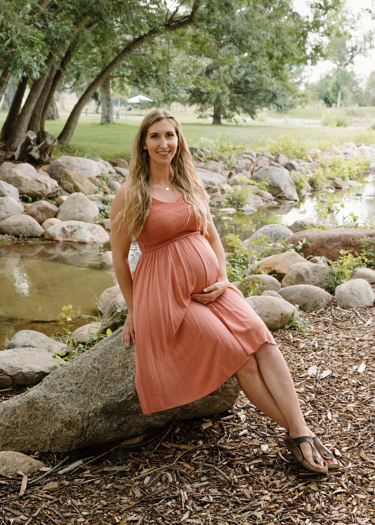 Pregnant woman in coral dress posing by rocky stream in lush park, cradling baby bump during outdoor maternity photo session.