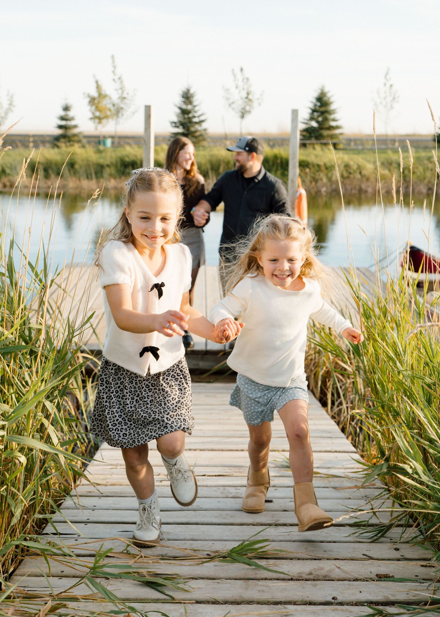 Two little girls running on wooden dock while parents walk behind near a scenic pond.