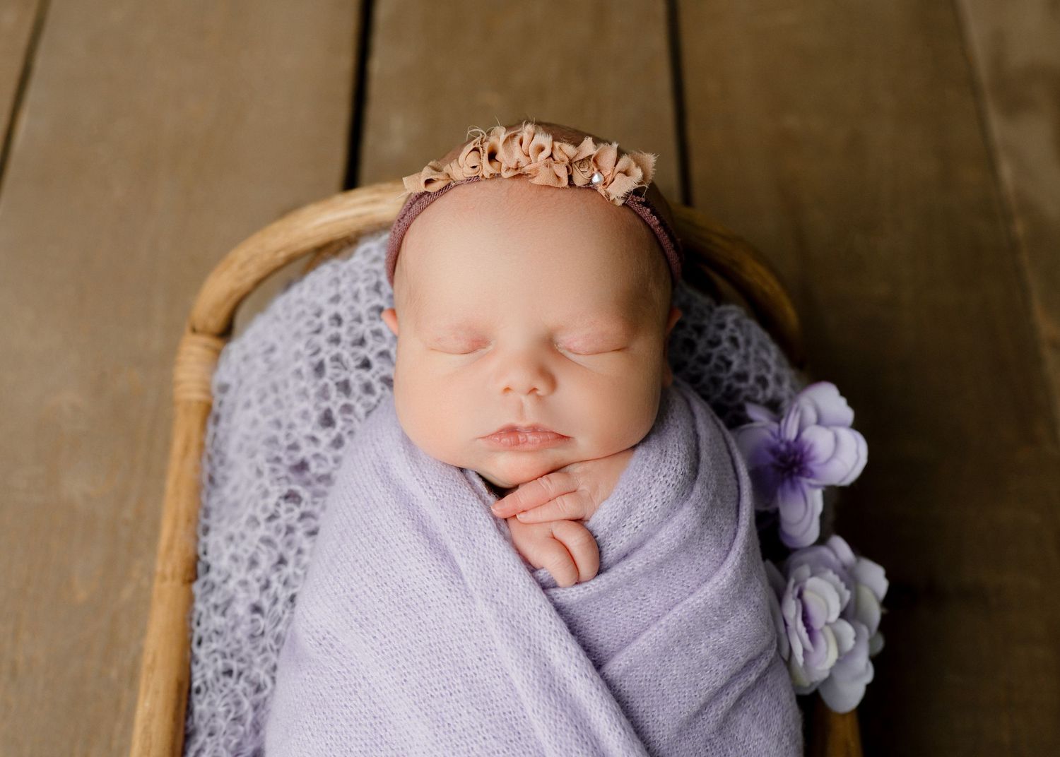 Sleeping newborn baby wrapped in lavender swaddle with pink floral headband in a wicker basket.