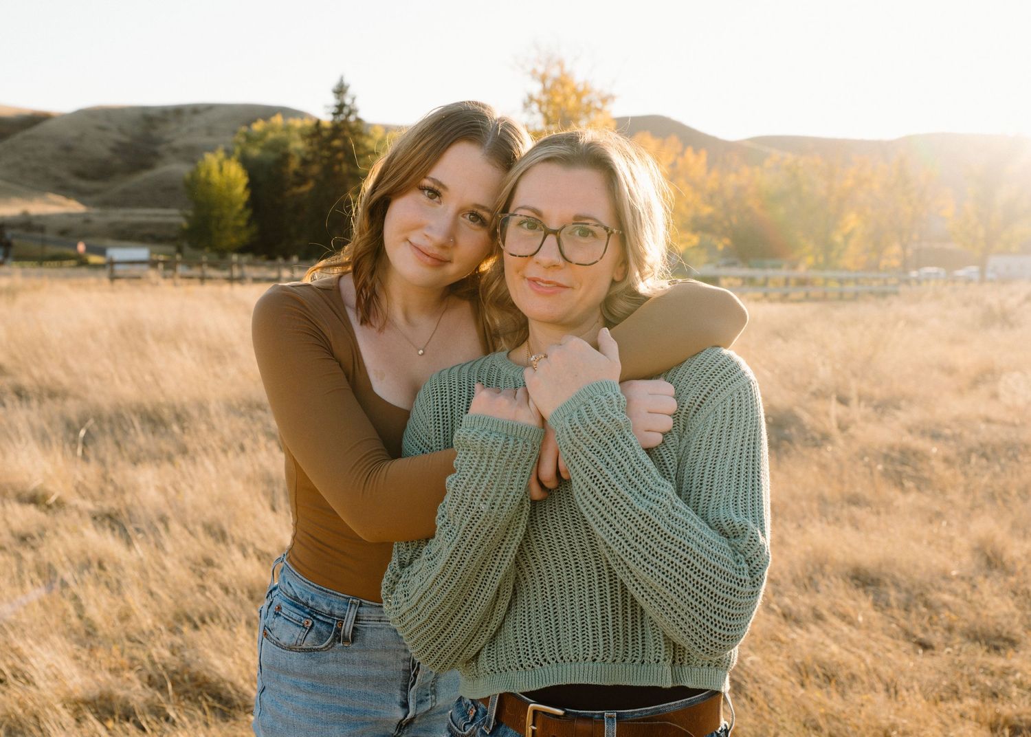 Two young women embrace warmly in a golden field with rolling hills and autumn trees behind them.