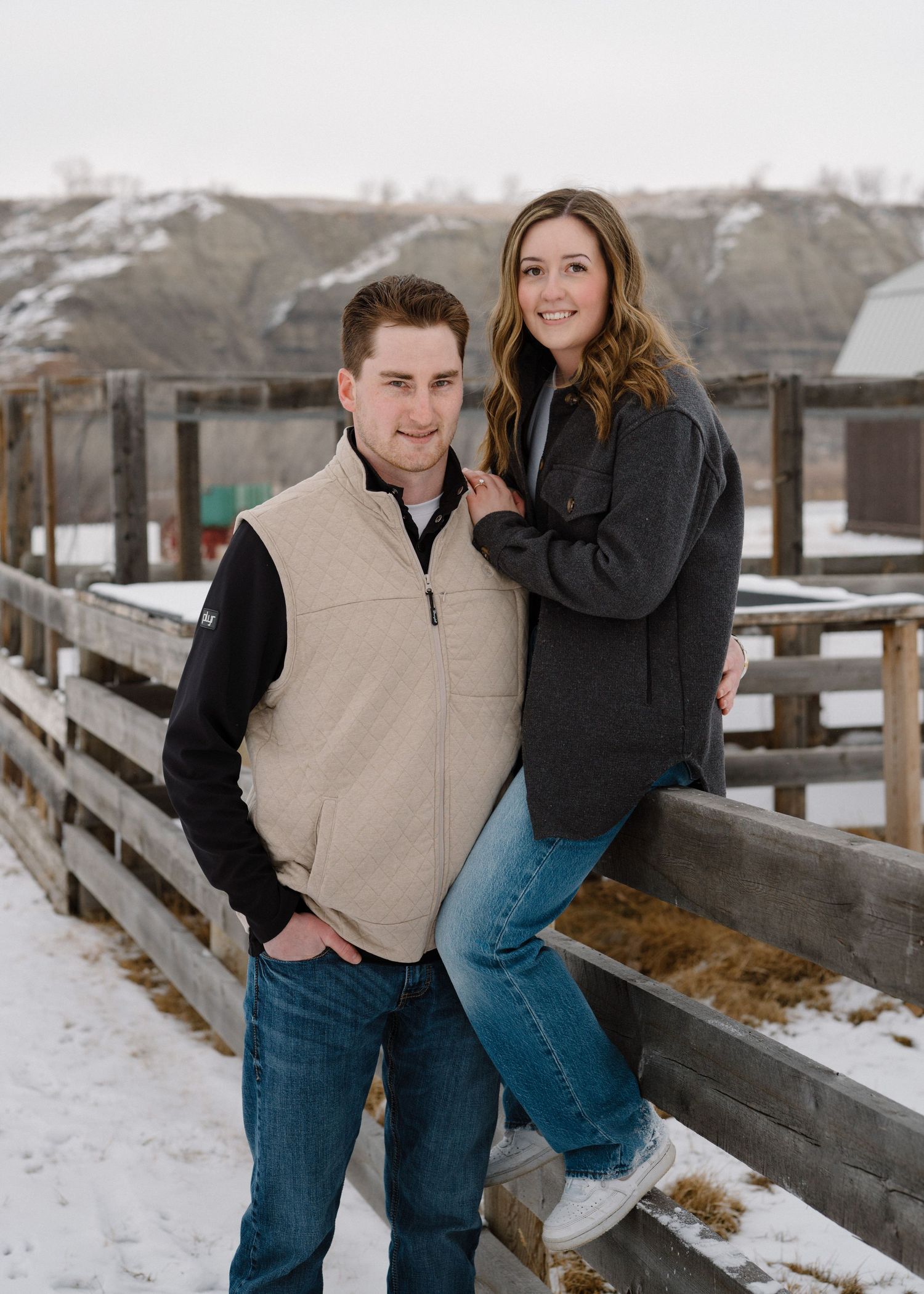 Engaged couple posing together on snowy farm fence in winter, woman leaning on mans shoulder.