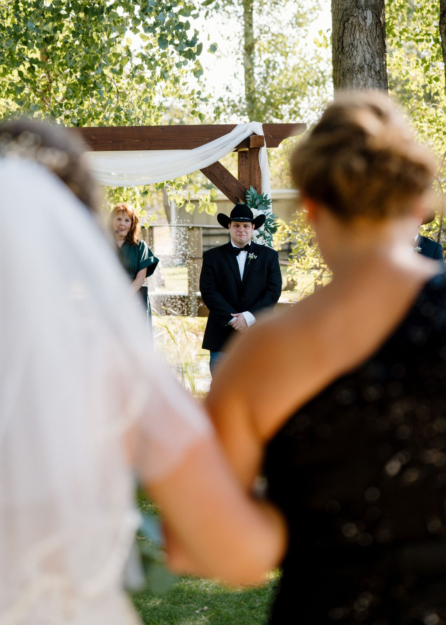 Groom in black tuxedo and cowboy hat waits at altar watching bride walk down the aisle.