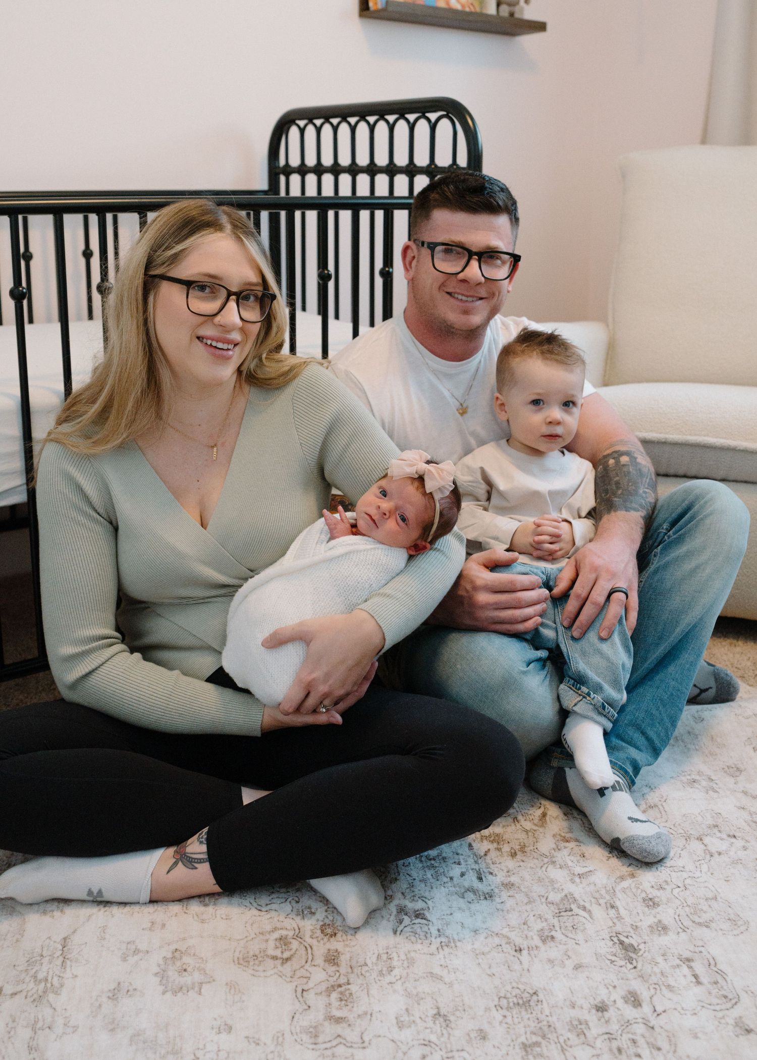 Family of four with newborn baby girl and toddler sitting together in decorated nursery.