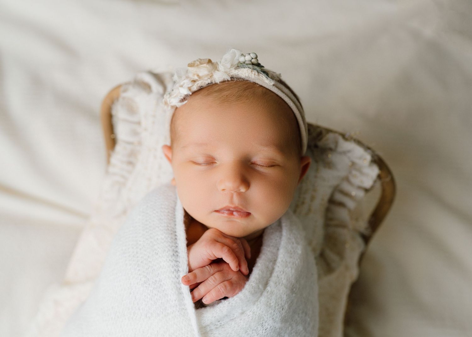 Peaceful newborn baby sleeping swaddled in white wrap wearing a delicate floral headband in a wicker basket.