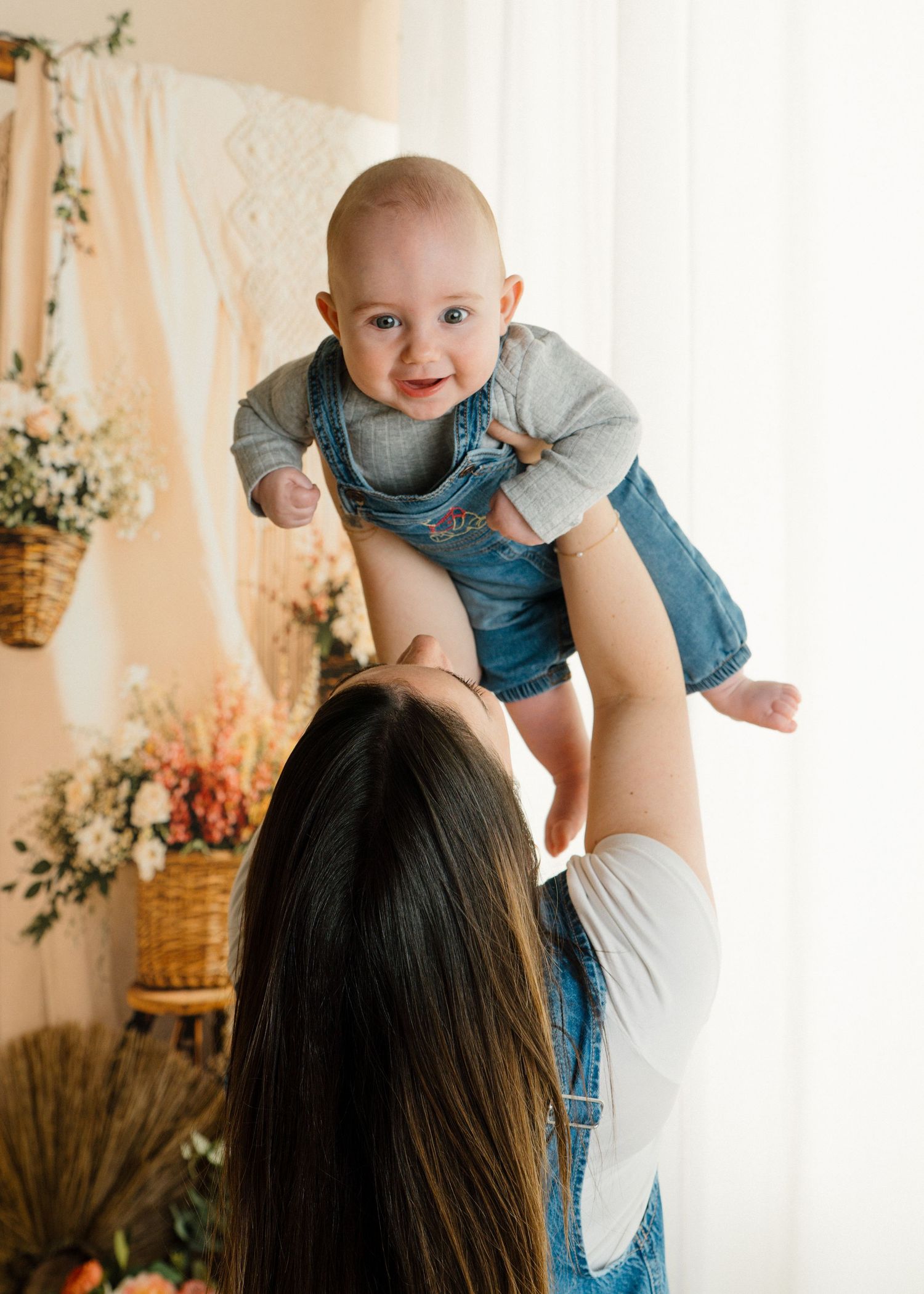Smiling baby in denim overalls lifted up by mother in a boho-styled photography studio.