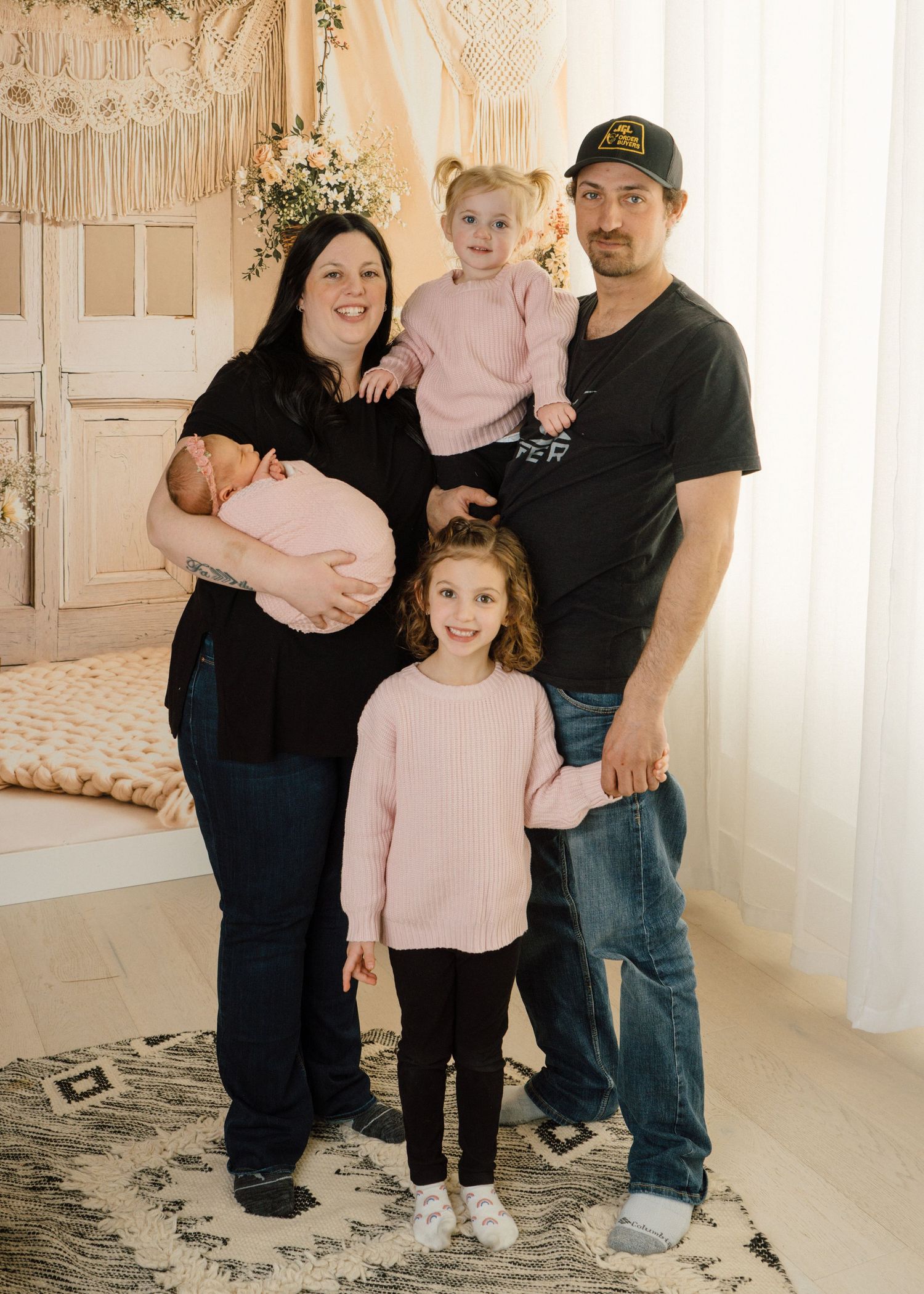 Family of five in black and pink outfits pose with newborn baby in a bright studio setting.
