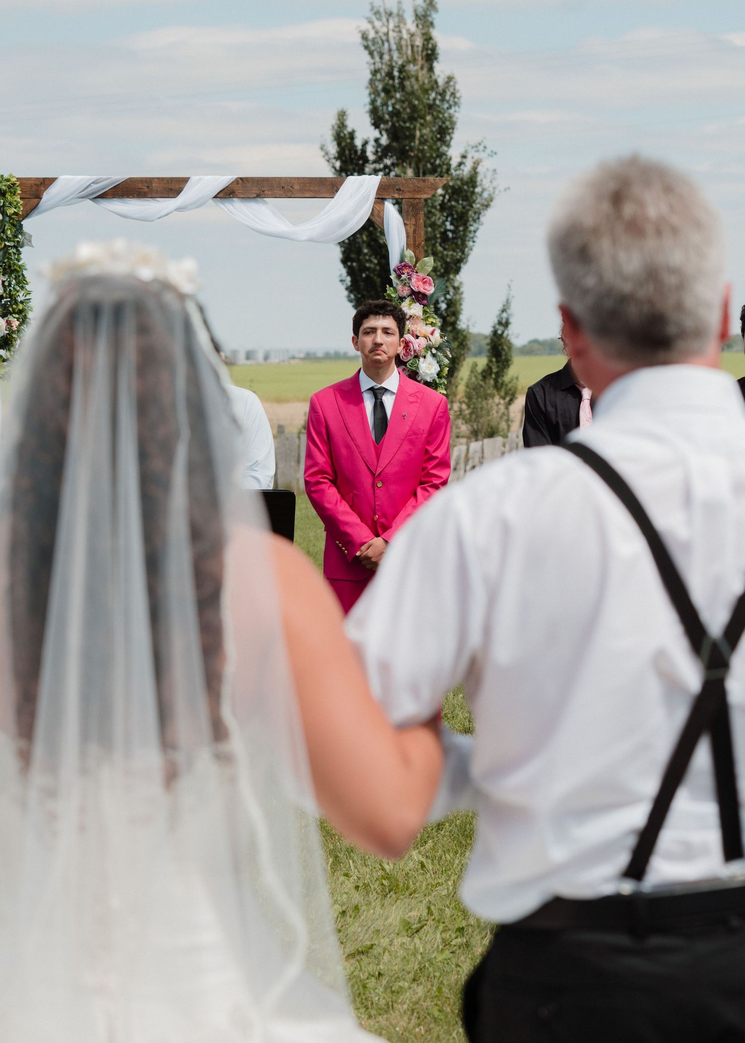 Groom in pink suit watches bride walk down aisle at outdoor wedding ceremony.