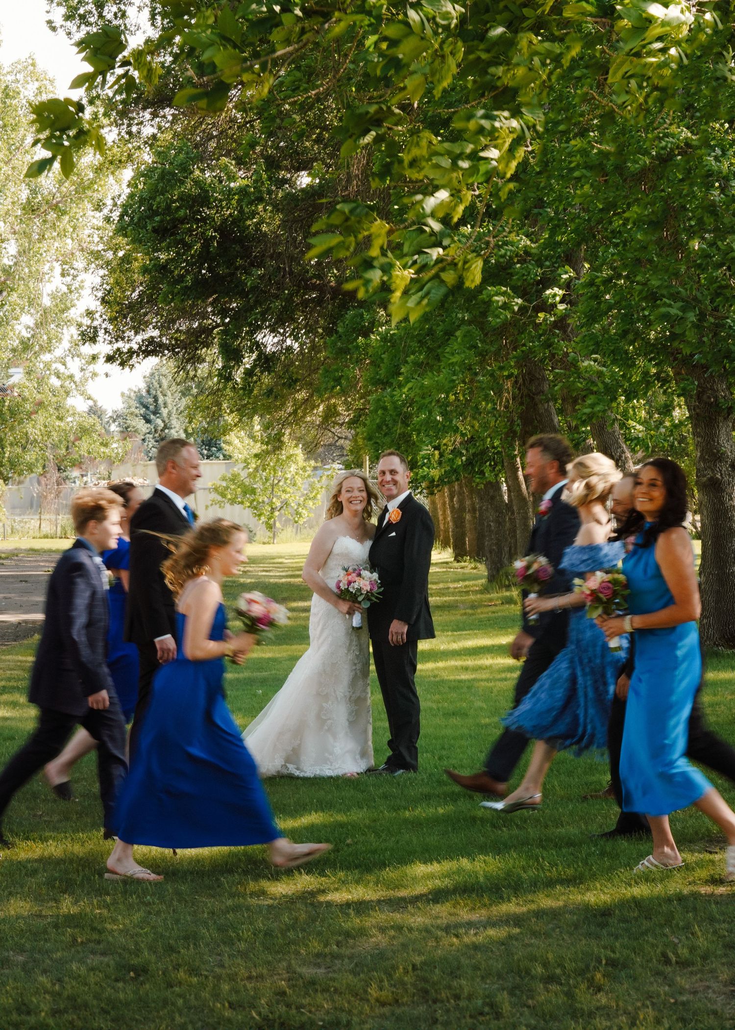 Wedding party in blue dresses and dark suits walks through a sunlit tree-lined outdoor venue.